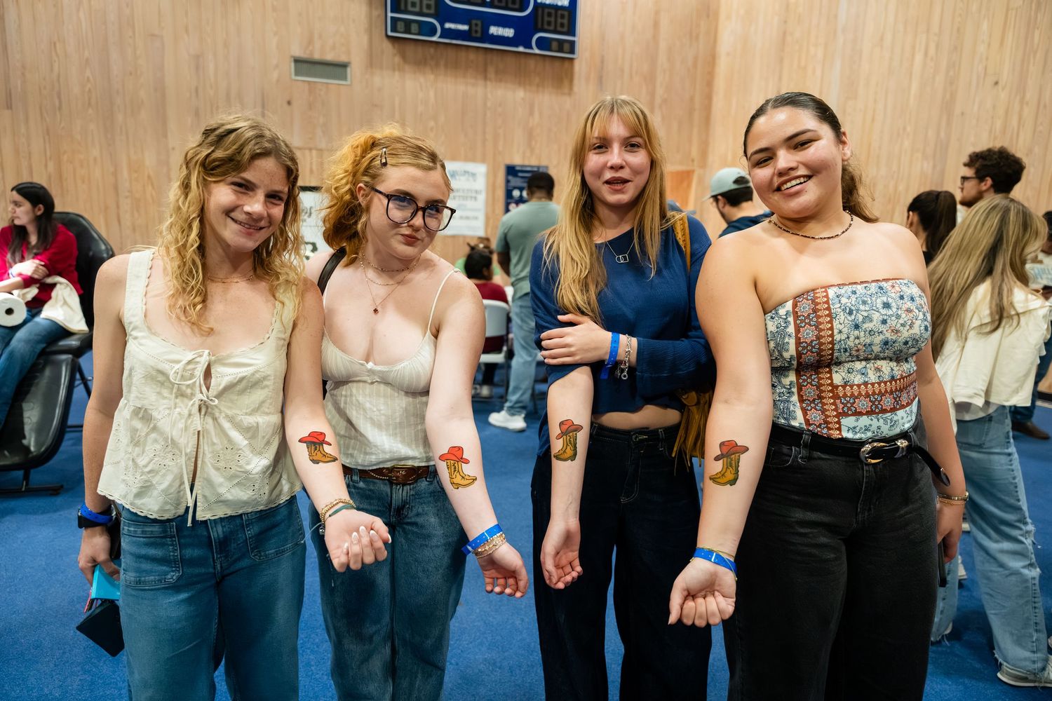 Four young women smiling and showing off matching temporary tattoos on their forearms at an indoor event.