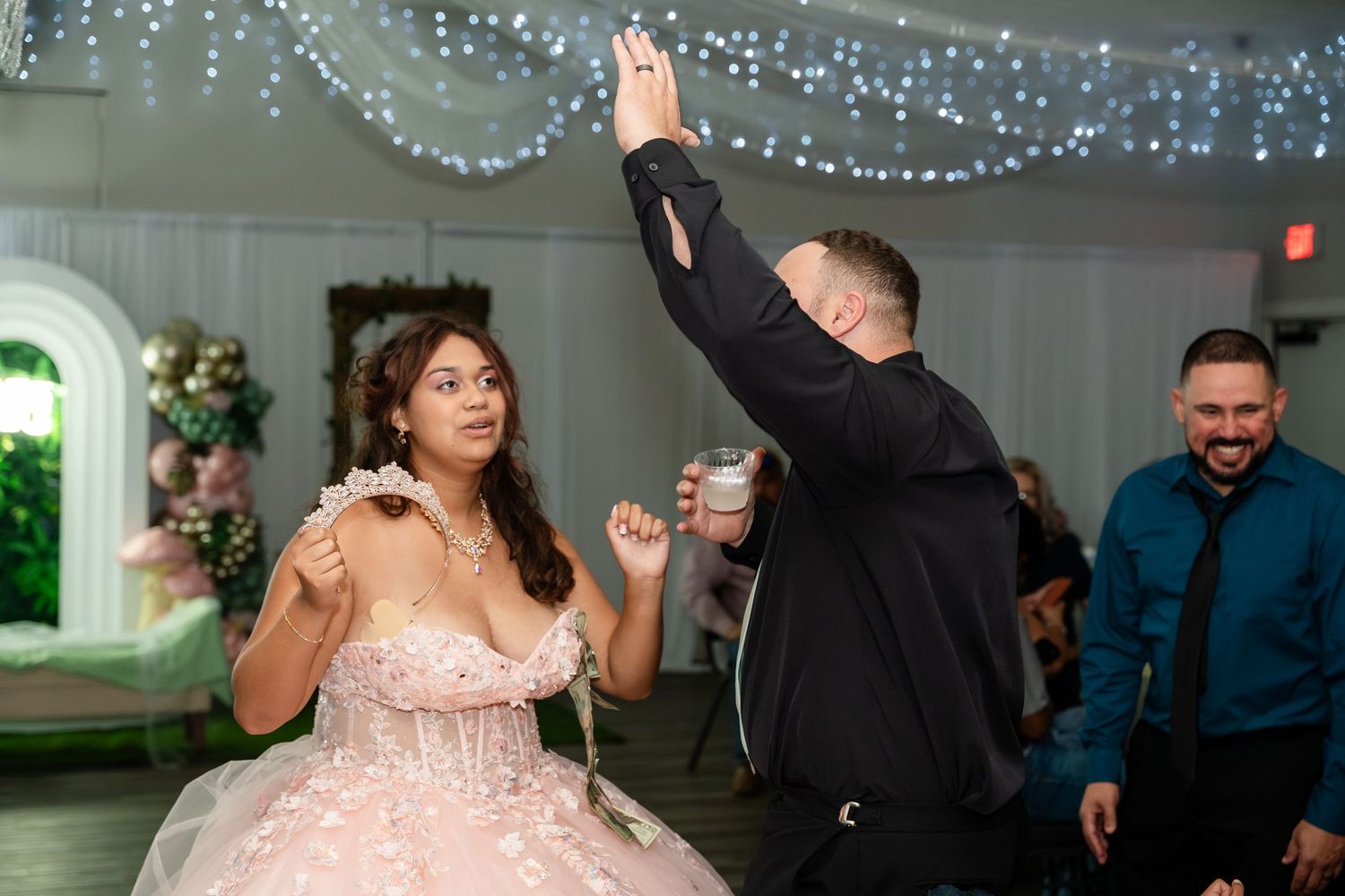 Young woman in pink ballgown dancing with man at formal celebration event with decorative lighting.
