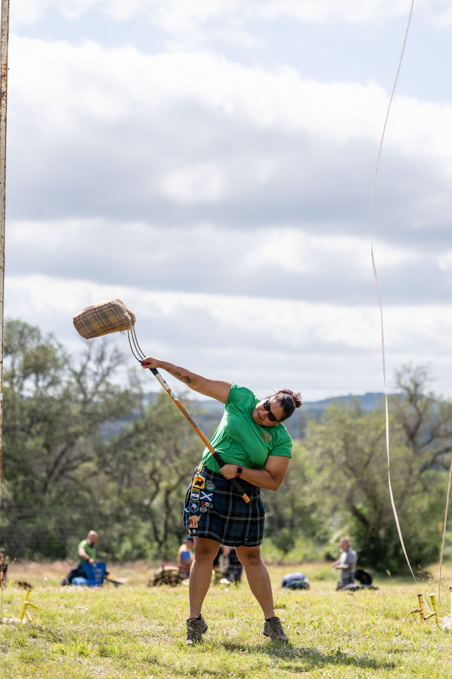 Man in green shirt and kilt performing Highland Games hammer throw on grassy field at outdoor Scottish festival.