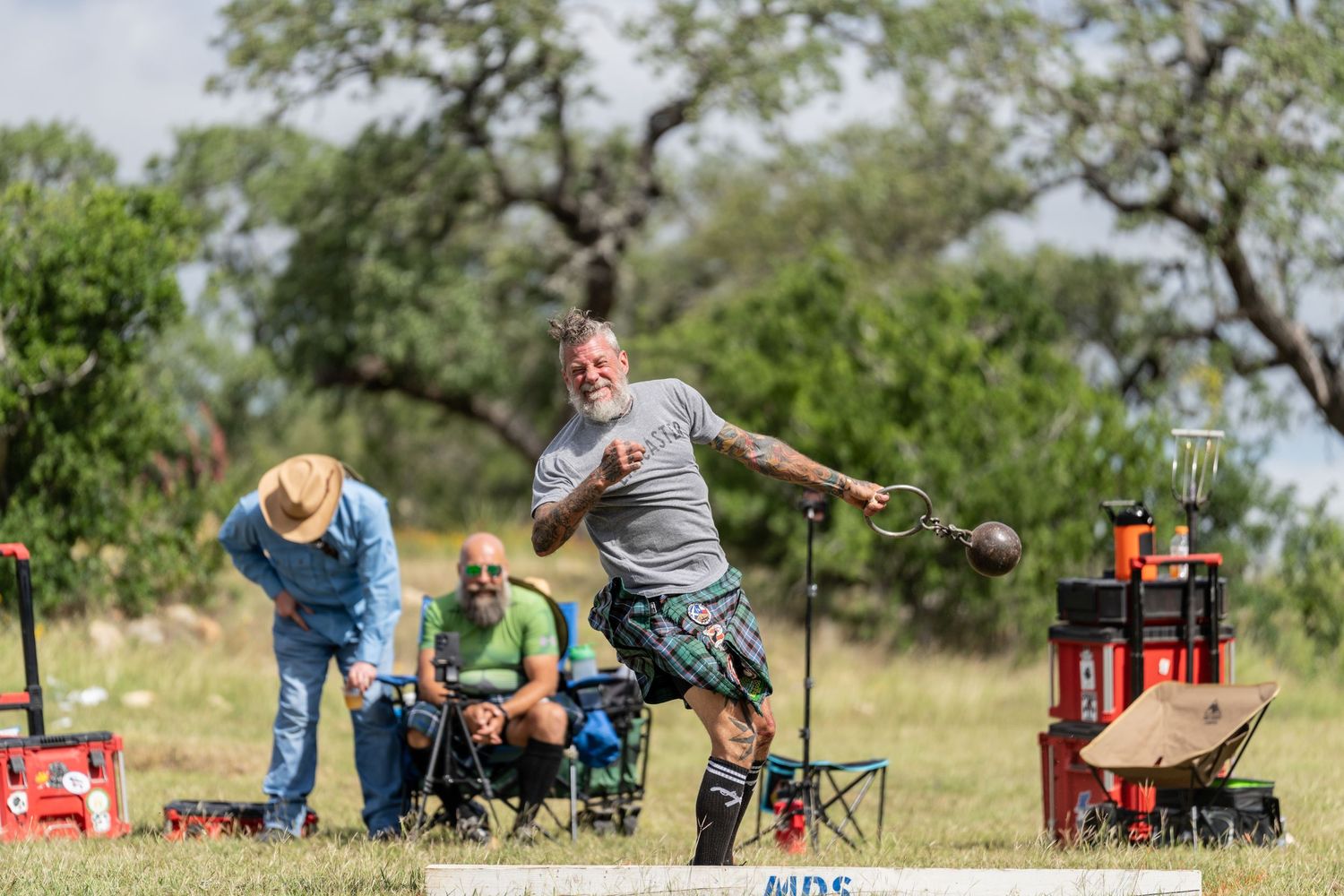 Bearded man in kilt throws heavy weight at Highland Games competition outdoors with spectators watching nearby.