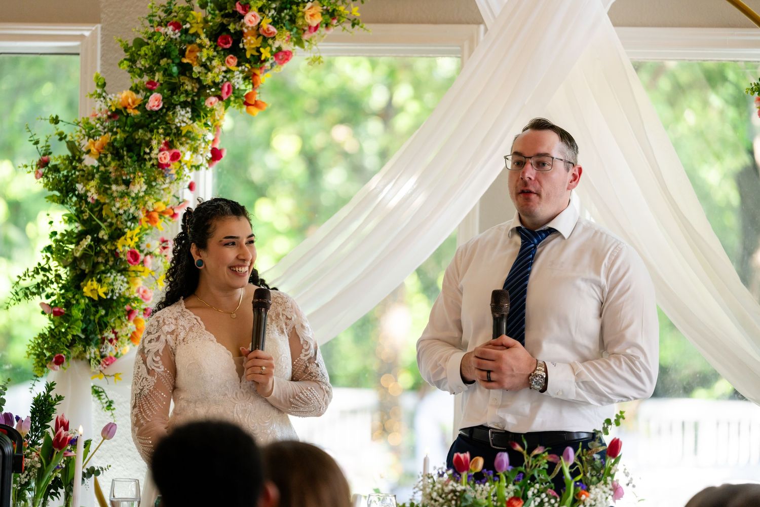 Bride and groom smiling during wedding reception speech beside floral arch with colorful flowers and white draping.