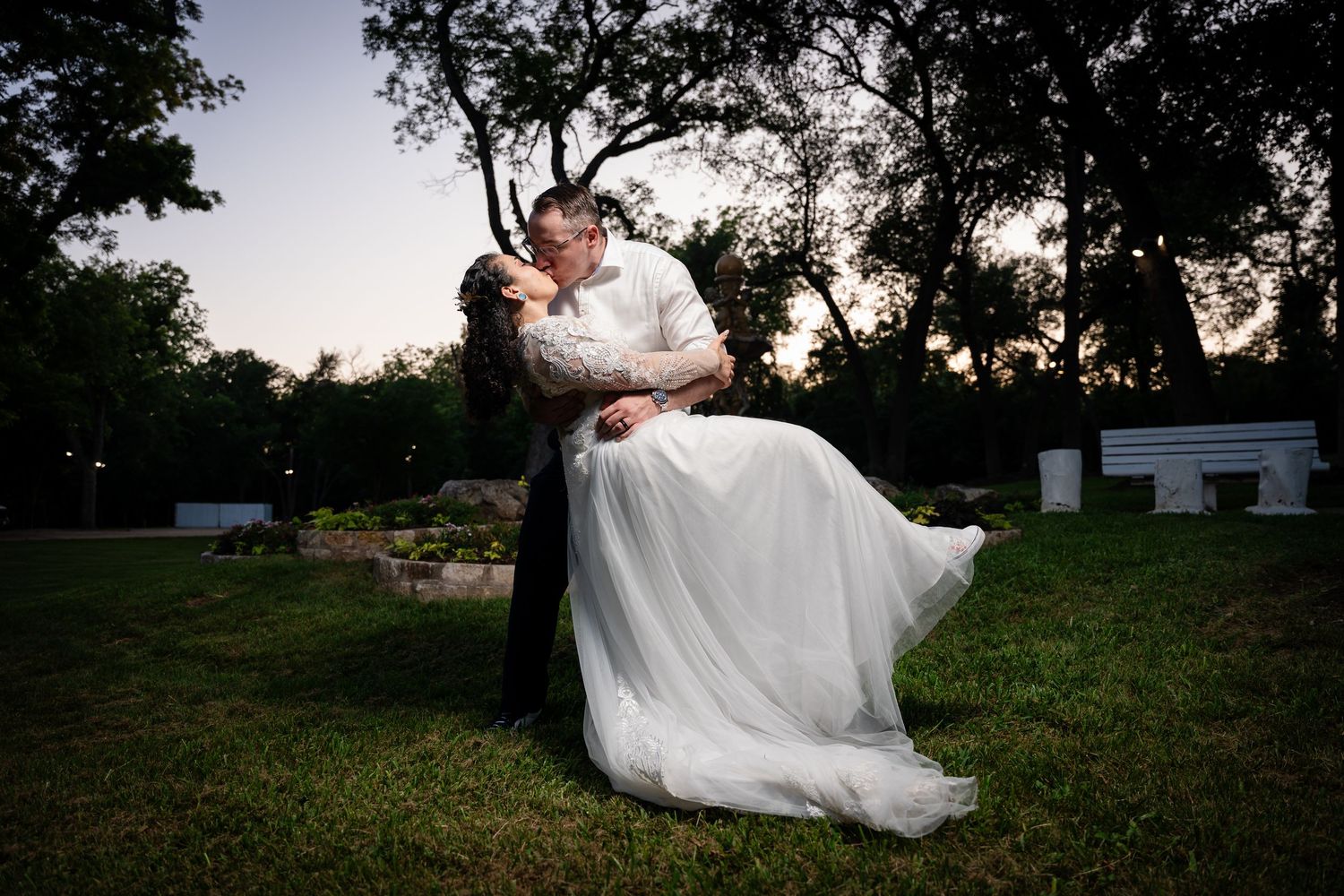 Groom lifts bride in white gown for romantic dusk kiss outdoors, trees and soft evening sky in background.