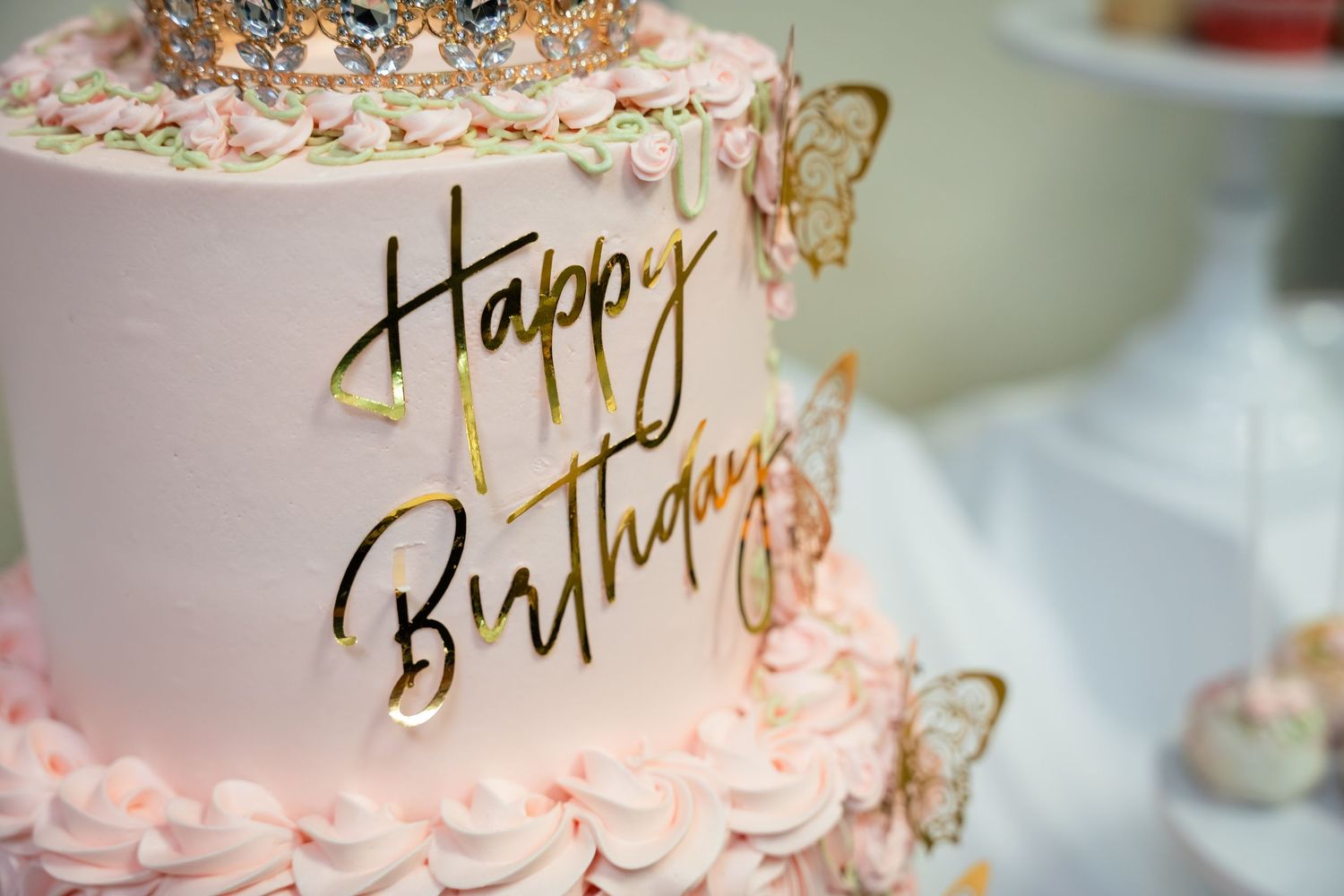 Elegant two-tier pink birthday cake with gold Happy Birthday topper, rosette frosting, and butterfly decorations.