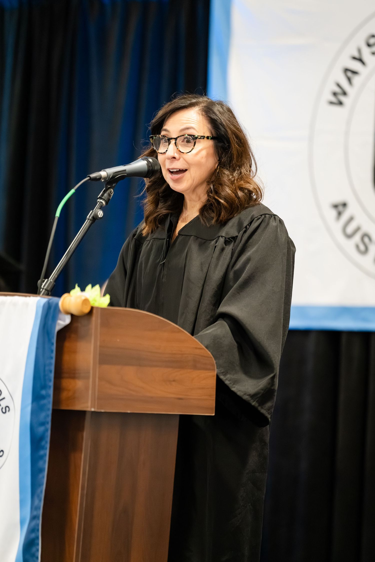 Woman in graduation gown speaking at podium with microphone during a graduation ceremony.