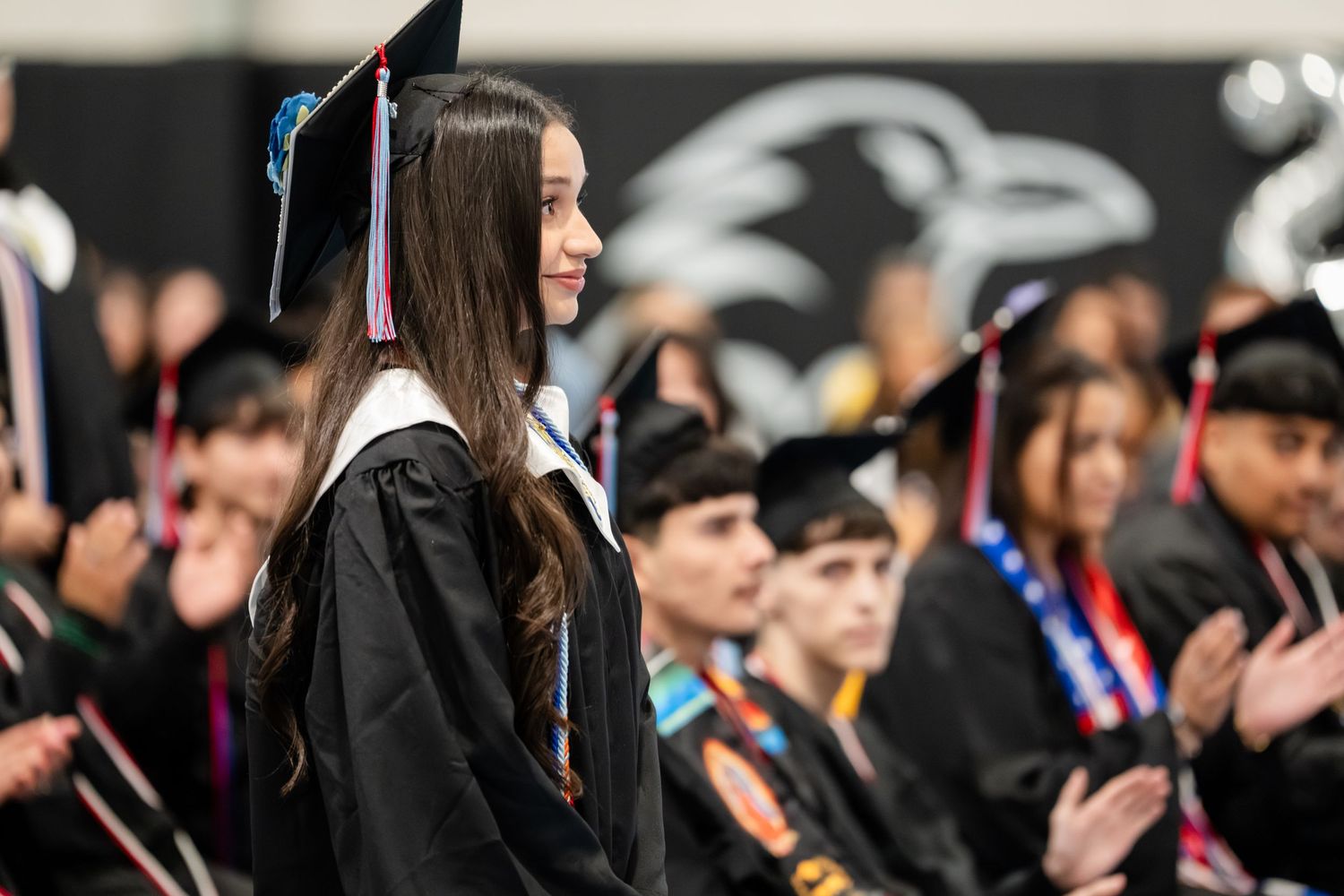 Student in graduation cap and gown smiles during commencement ceremony with classmates seated in background.