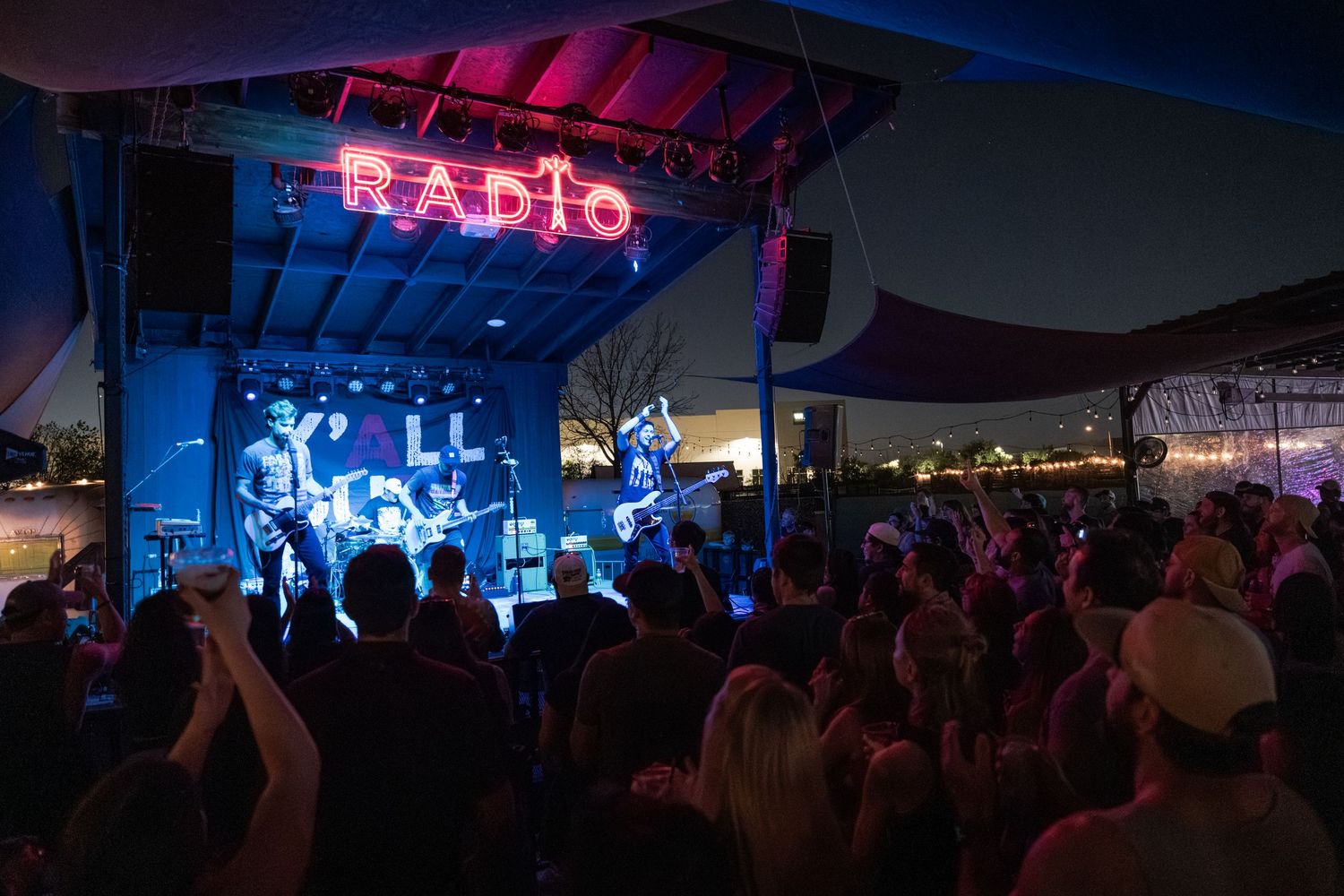 Crowd watching a live band perform on an outdoor stage at night under a glowing neon Radio sign.