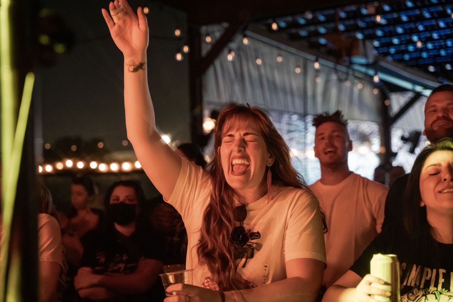 Excited woman raises hand at outdoor concert venue at night, crowd behind her, string lights glowing.