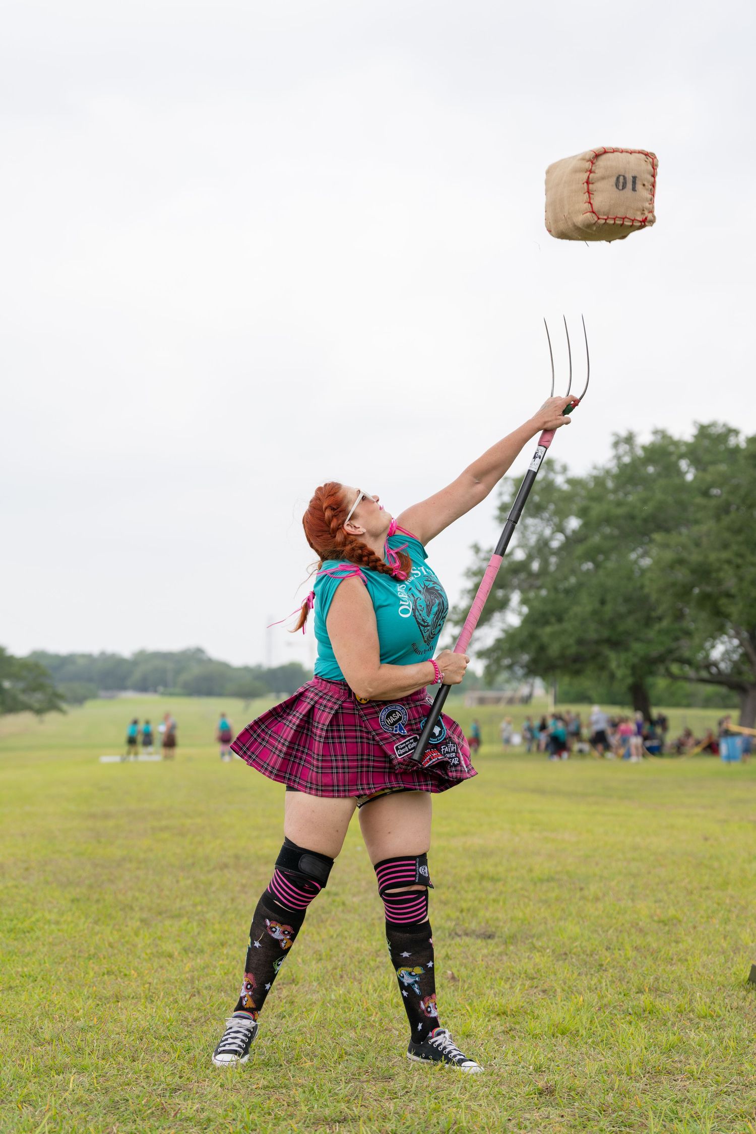 Woman in plaid skirt and teal top tosses a weighted bag on a pitchfork at an outdoor Highland Games event.