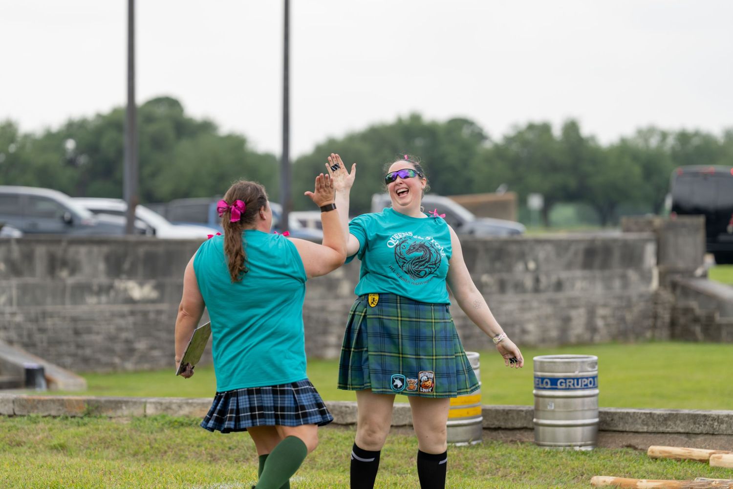 Two women in tartan kilts and teal shirts high-five at a Scottish Highland Games outdoor event.