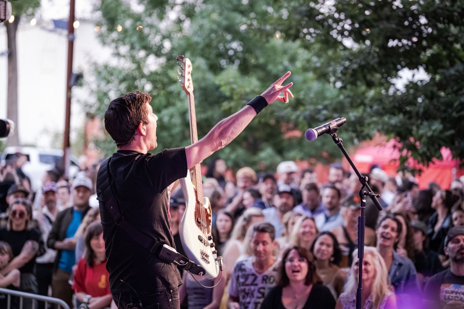 A rock musician performs on an outdoor stage, raising his hand to an enthusiastic crowd at a lively concert event.