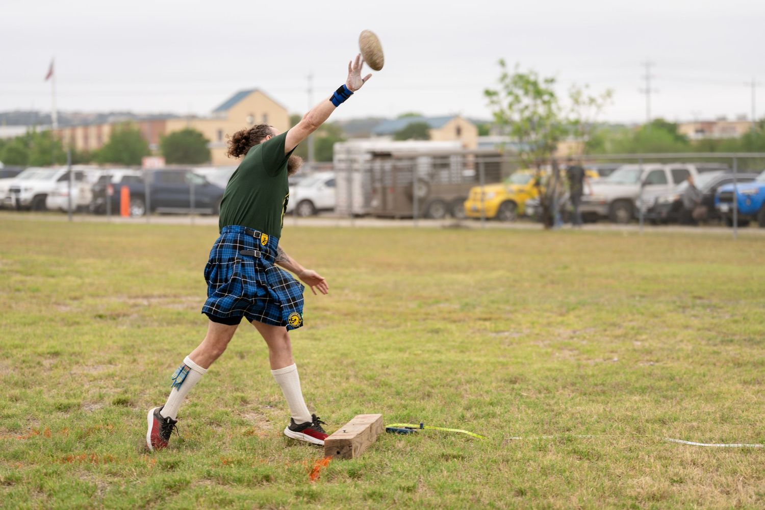 Man in tartan kilt throwing a weight at a Highland Games competition on a green field.