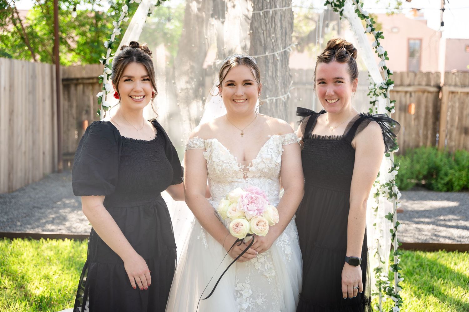 A wedding party poses outdoors with decorative floral arch and string lights.