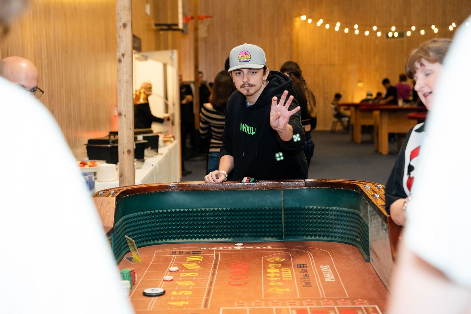 Players gathered around a green felt gaming table with string lights in background.