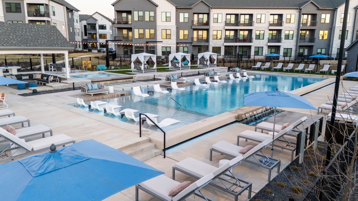 Modern apartment complex pool area featuring white lounge chairs and a spacious deck surrounded by residential buildings.