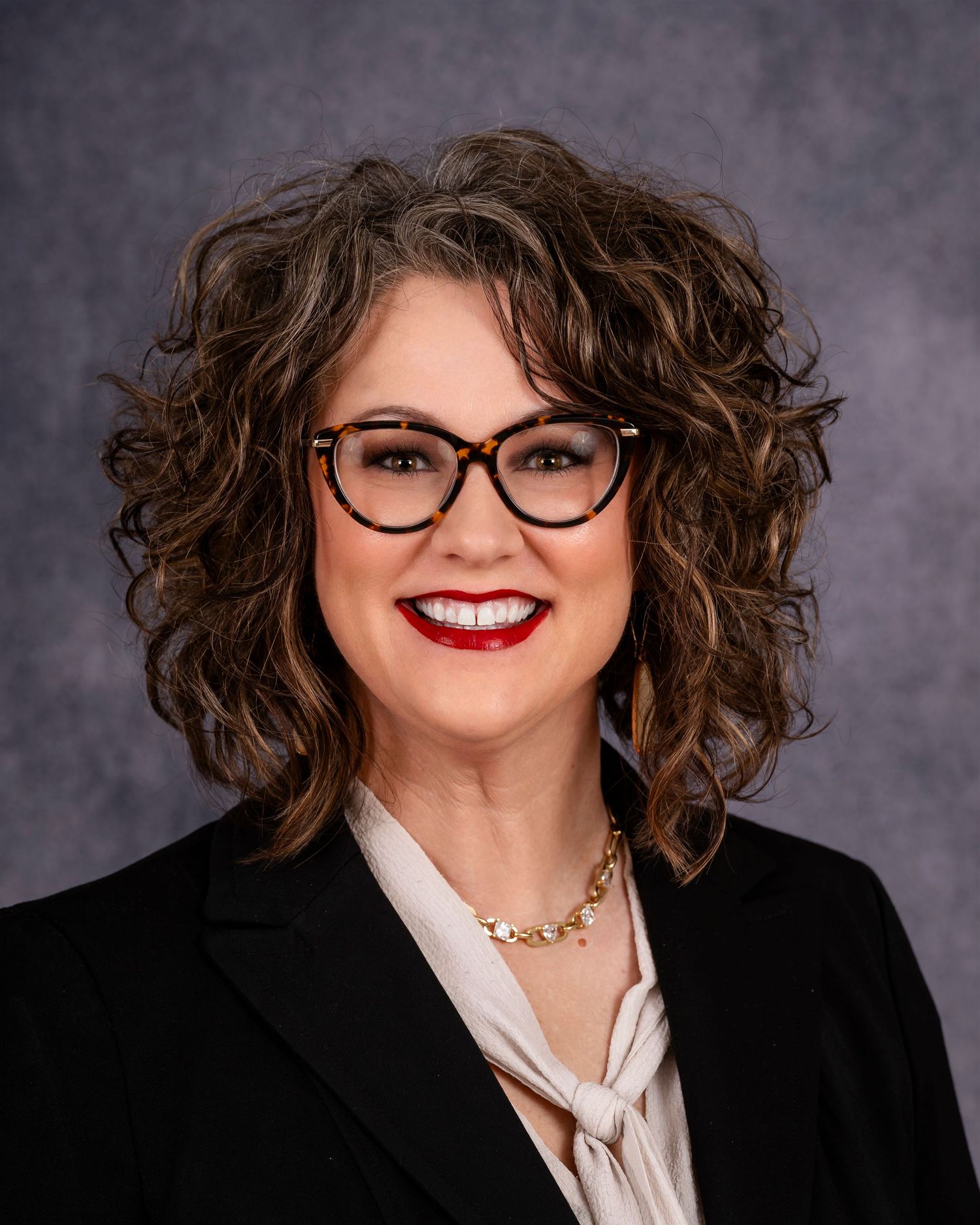 Professional headshot of someone wearing black-framed glasses, a blazer, white blouse and necklace with styled curly hair.