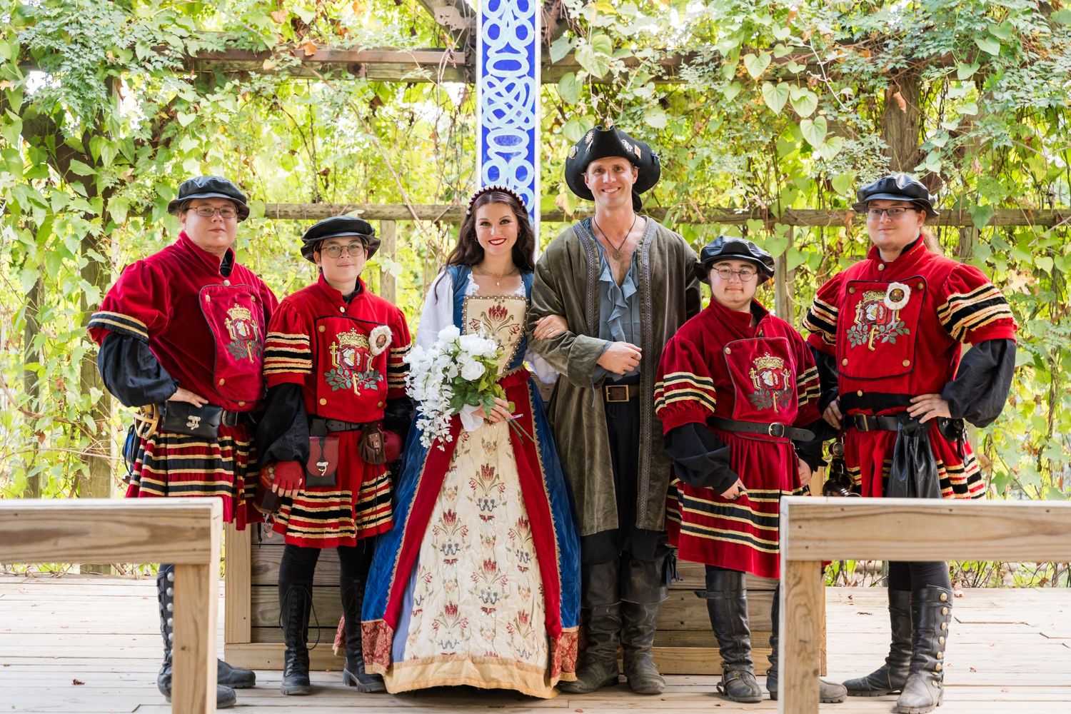 Wedding party poses with guards in red Tudor uniforms during renaissance fair celebration.
