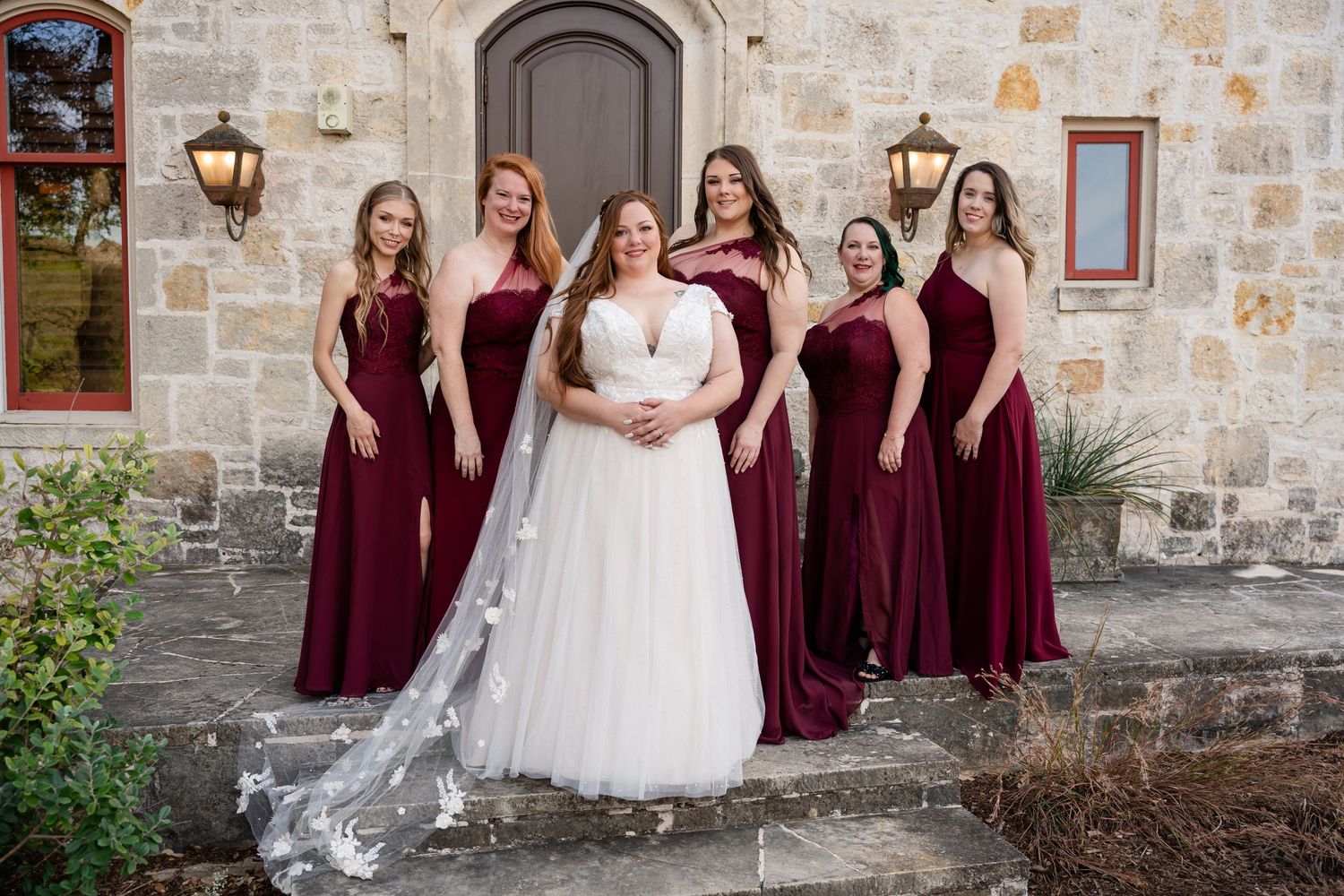 Wedding party in burgundy velvet dresses and white gown posed on stone steps.