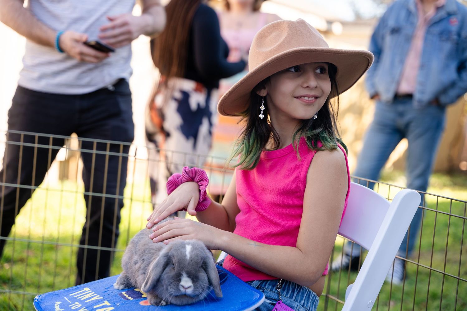 A young attendee in a pink outfit and brown hat holds a bunny at an outdoor event.