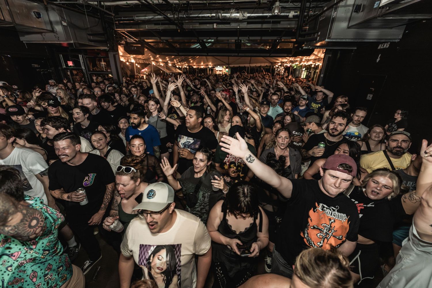 Energetic crowd at a rock concert with raised hands and illuminated venue ceiling.
