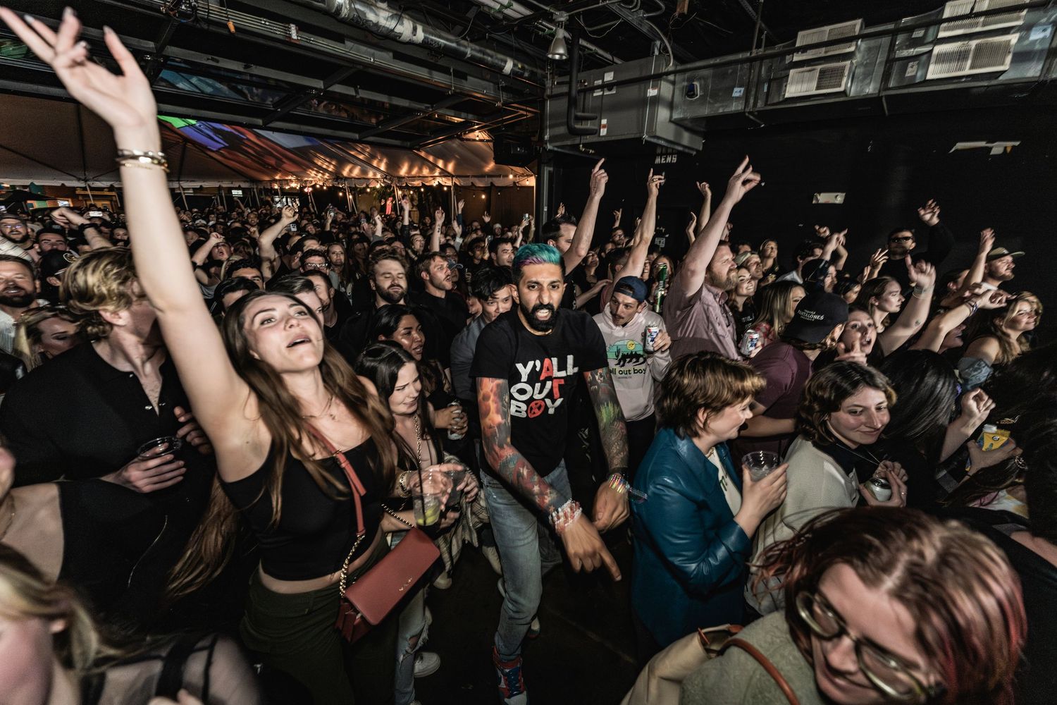 Energetic crowd raises their hands and cheers during a packed concert venue with dark lighting and industrial interior.