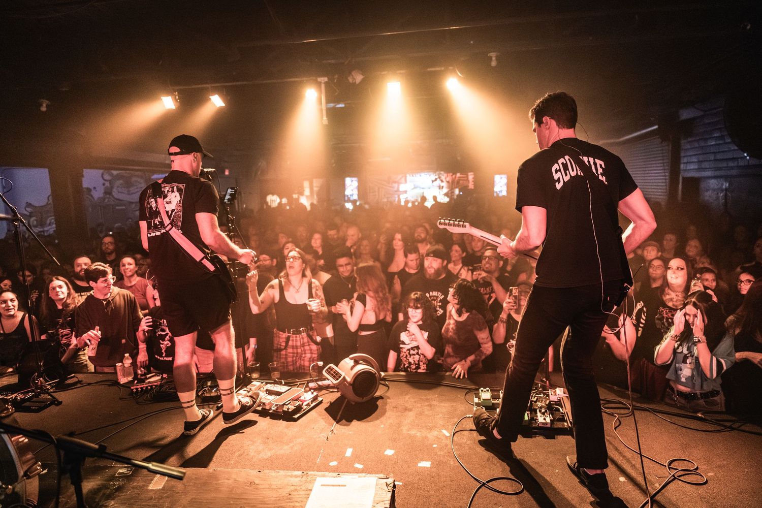 Concert performers on stage with silhouetted crowd and dramatic amber lighting at a small venue show.