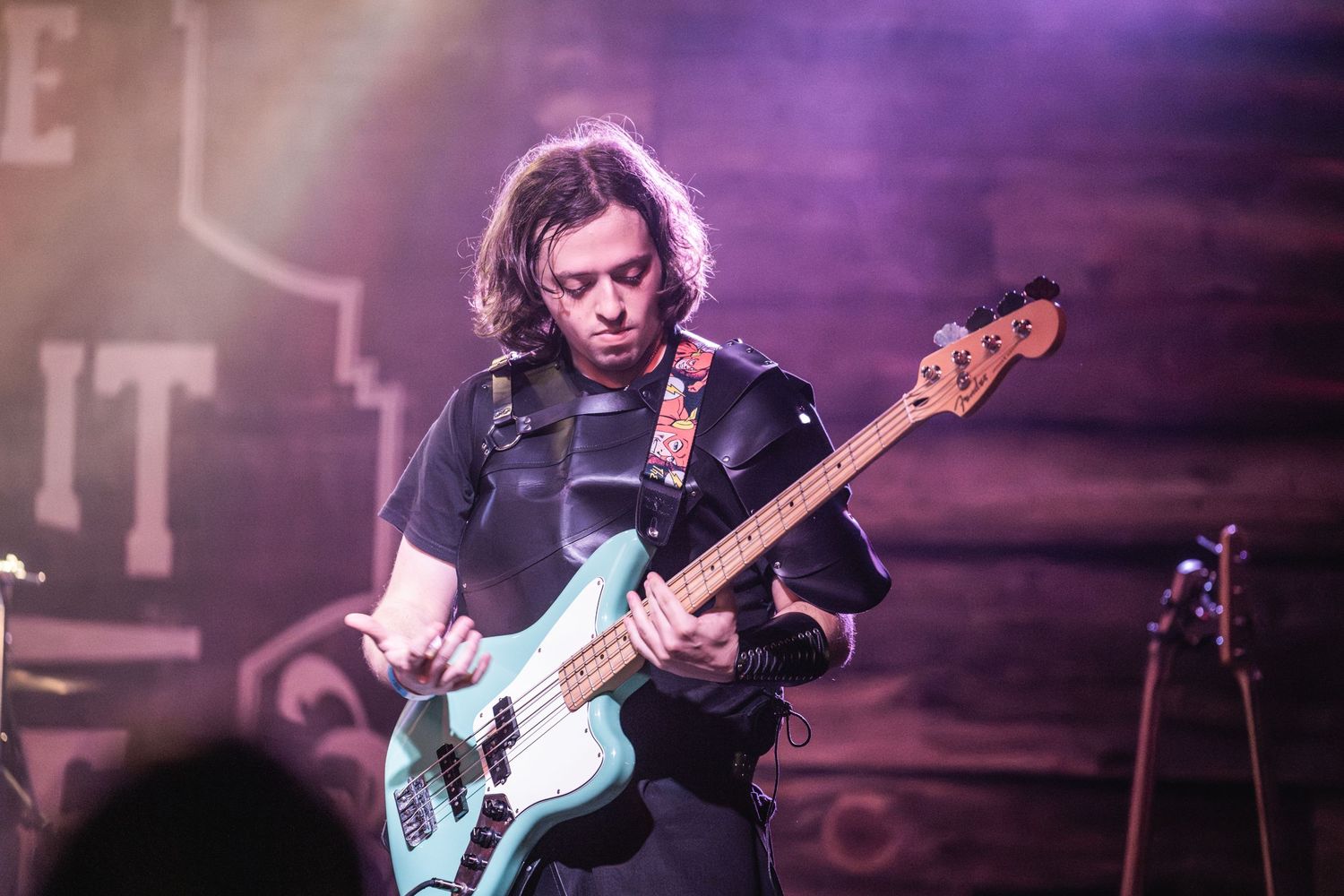 Bass guitarist plays a light blue Fender bass guitar under purple stage lighting during performance.