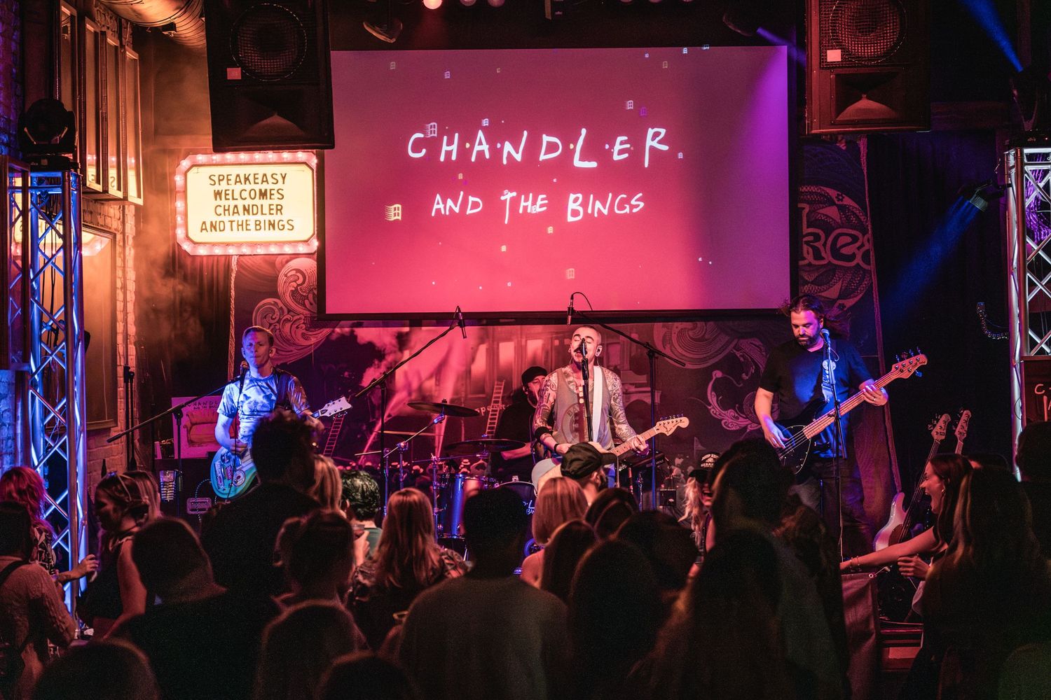 Rock band performs under Chandler projection screen with enthusiastic crowd at intimate venue.
