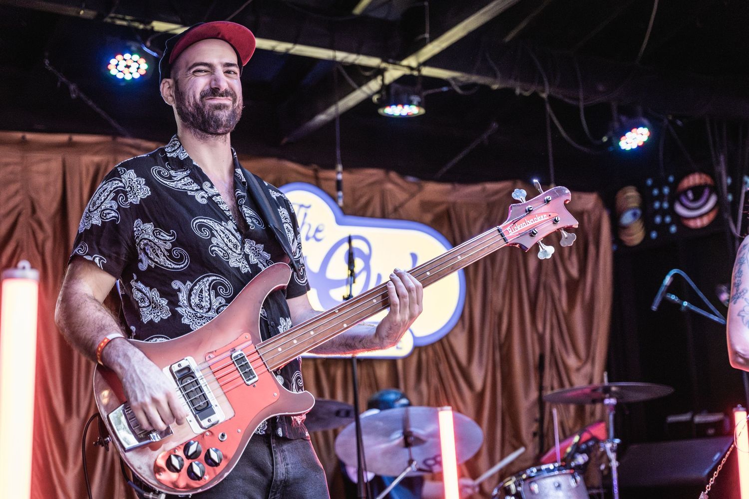 Bass guitarist in a black patterned shirt performs energetically on stage with lighting and decorative backdrop.