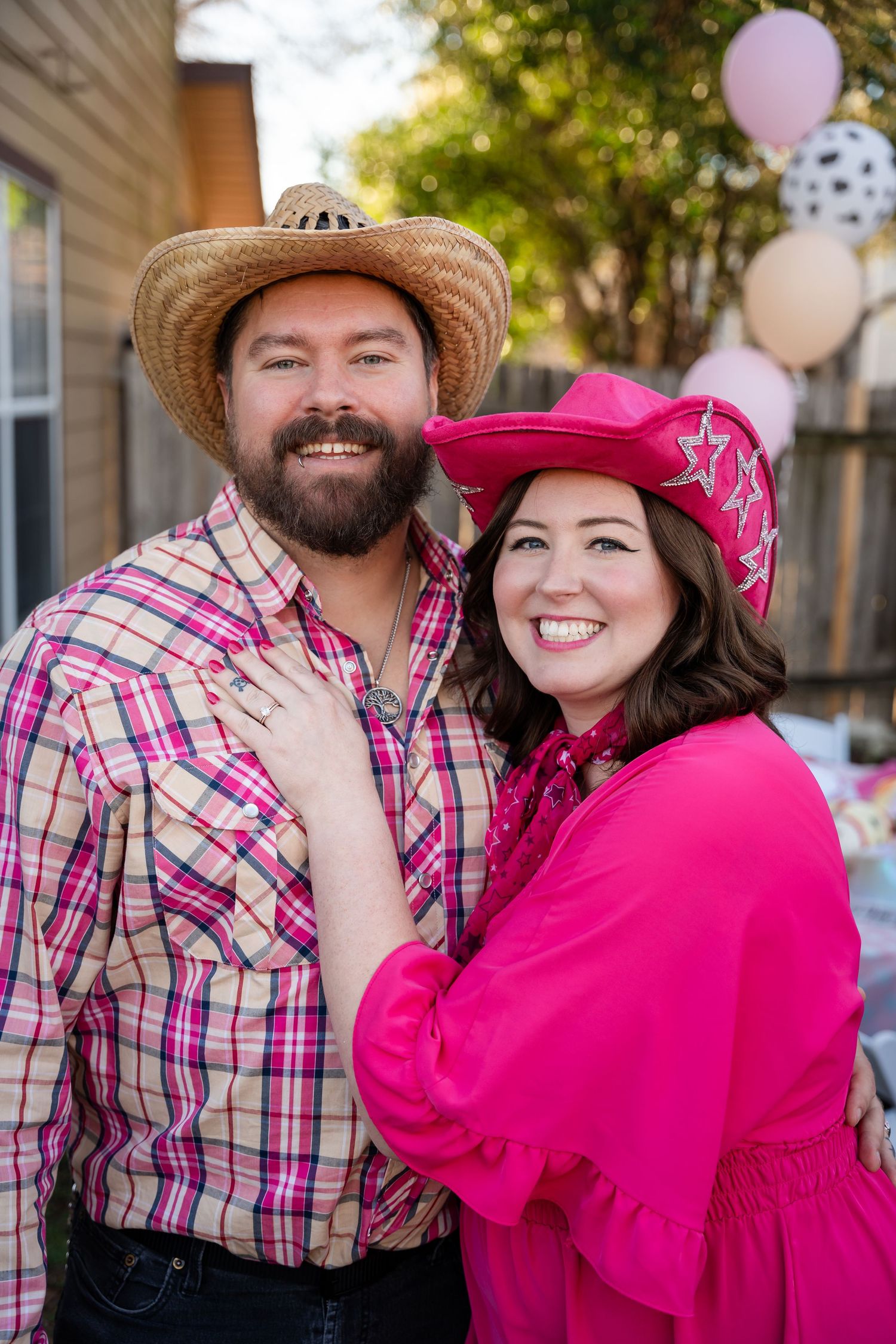 A couple wearing western hats and pink attire smile at an outdoor celebration with balloons.