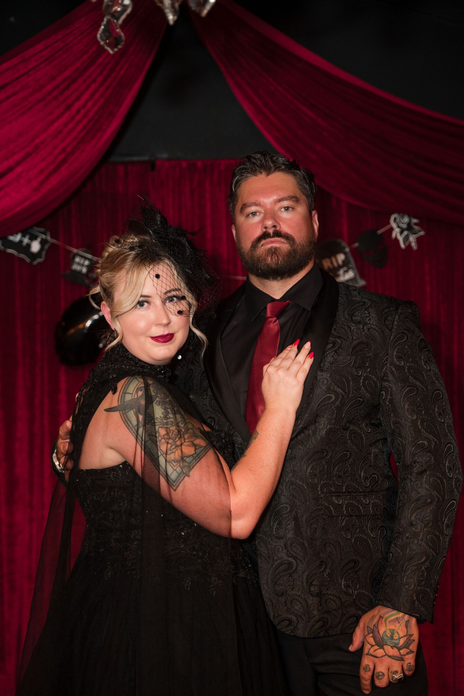 Formal couple in black attire pose against deep red draped backdrop.