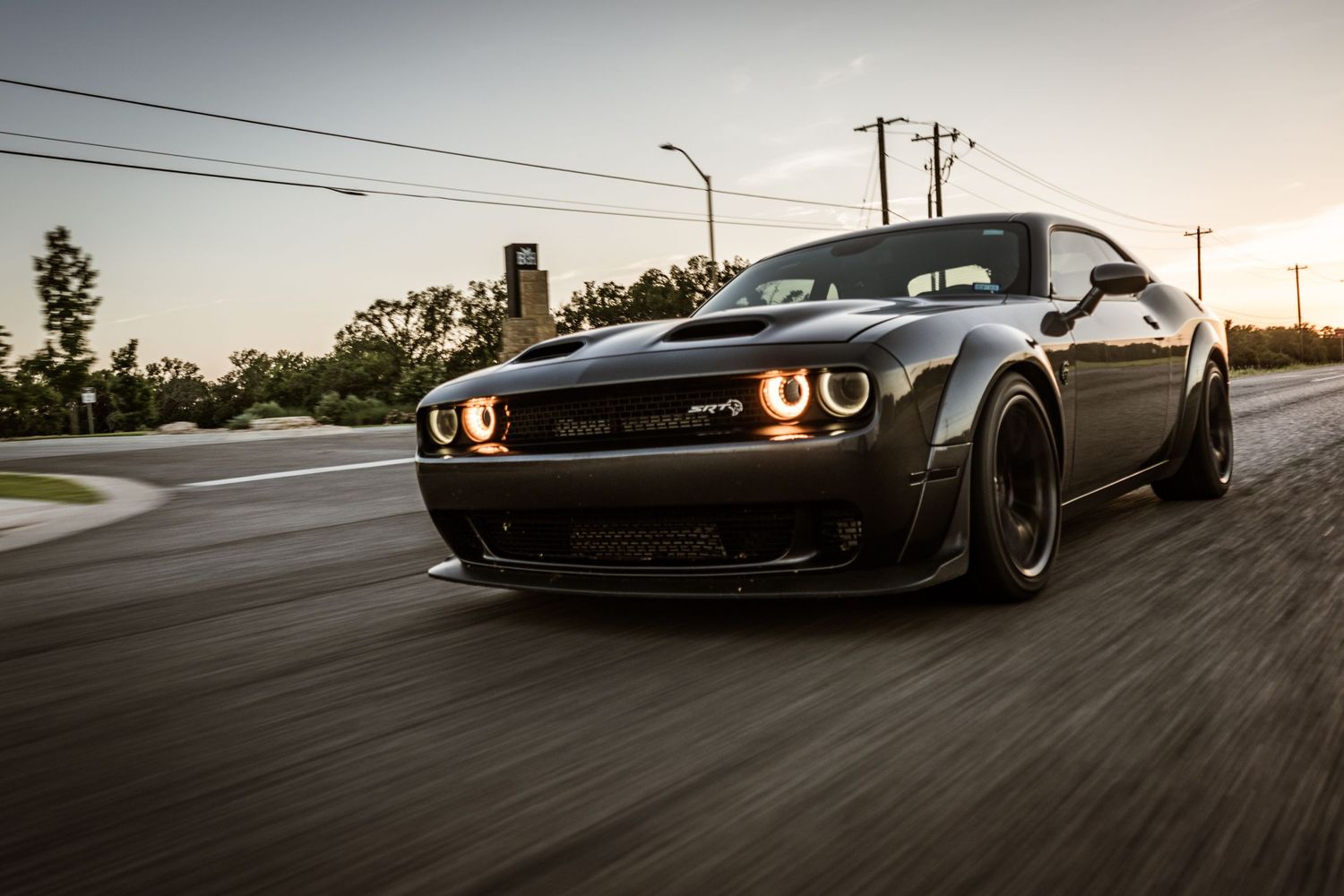 Matte black Dodge Challenger Demon with custom wheels driving on a curved road at sunset.
