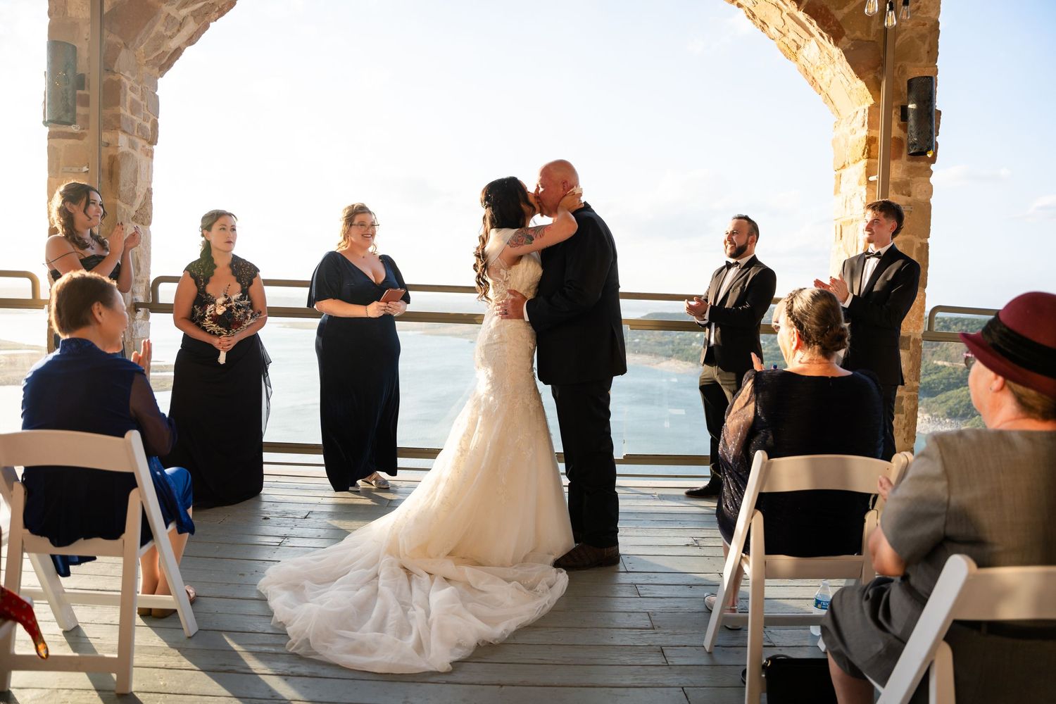 Newlyweds share first kiss during ceremony under stone arch with guests seated.