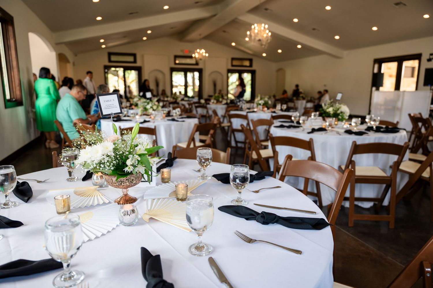 Wedding reception venue with round tables decorated in white linens and wooden folding chairs in an elegant dining space.