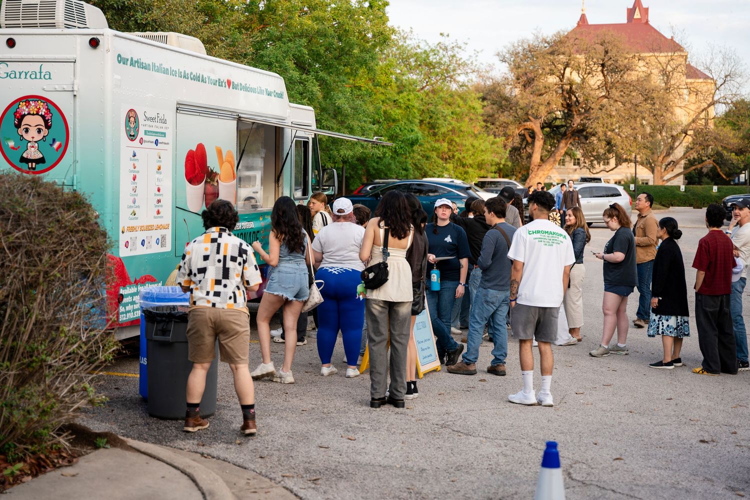 A group of customers waiting in line at a food truck on a sunny day with trees and buildings in the background.