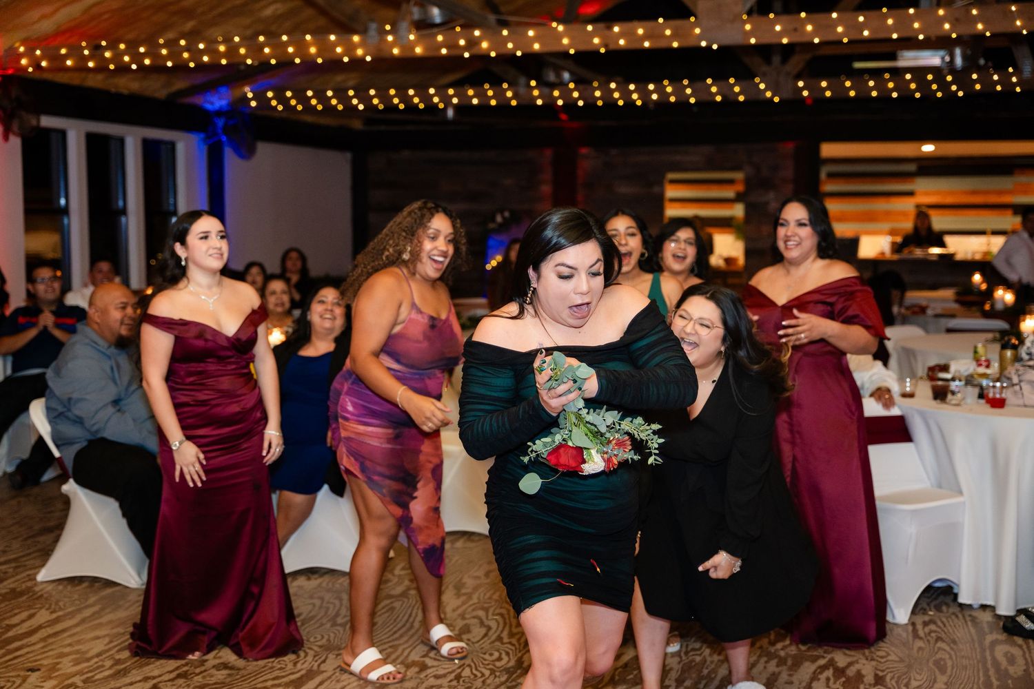 Wedding party members in burgundy dresses gather around a bouquet toss at the reception.