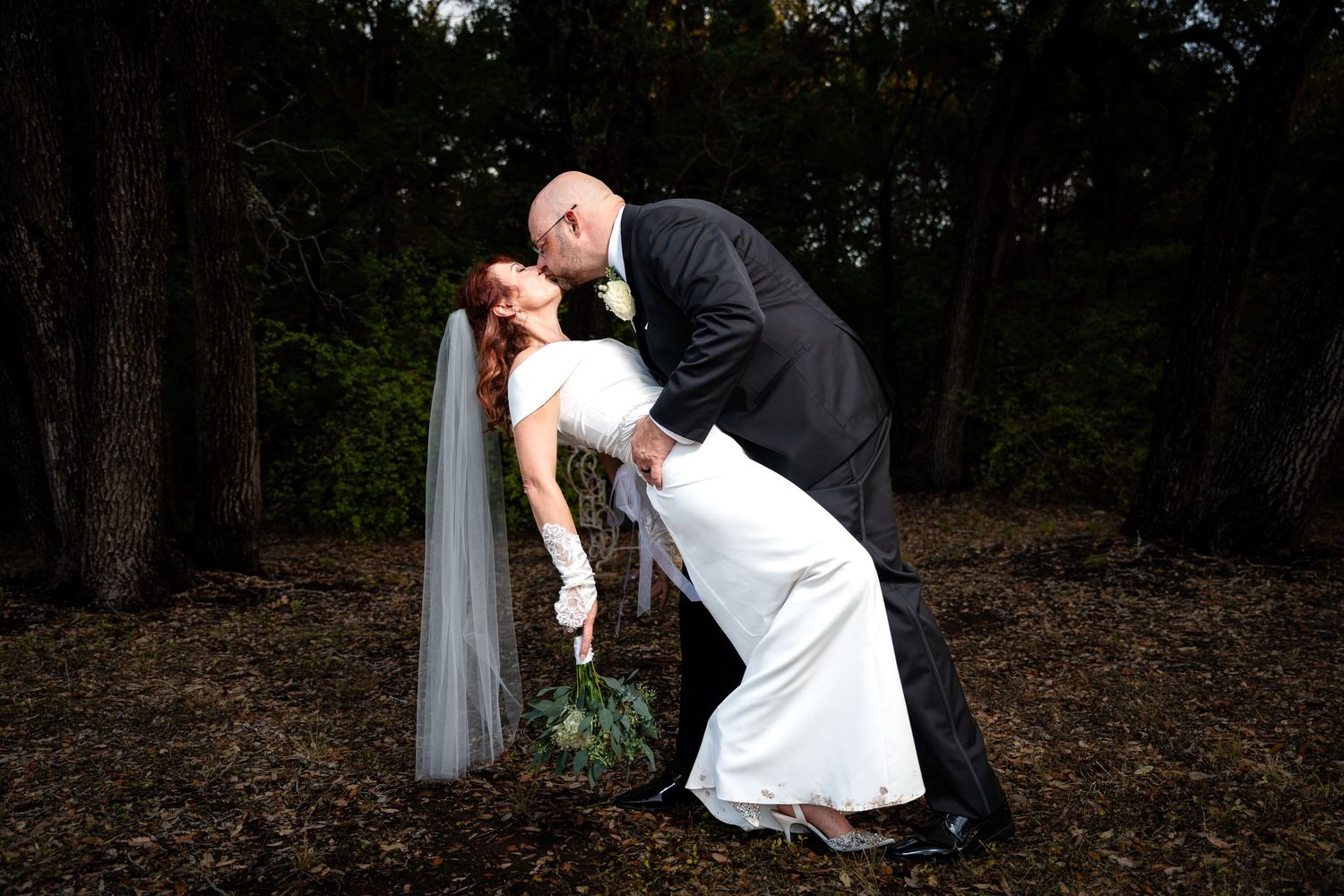 A romantic dip pose between a couple in wedding attire photographed in a wooded outdoor setting.