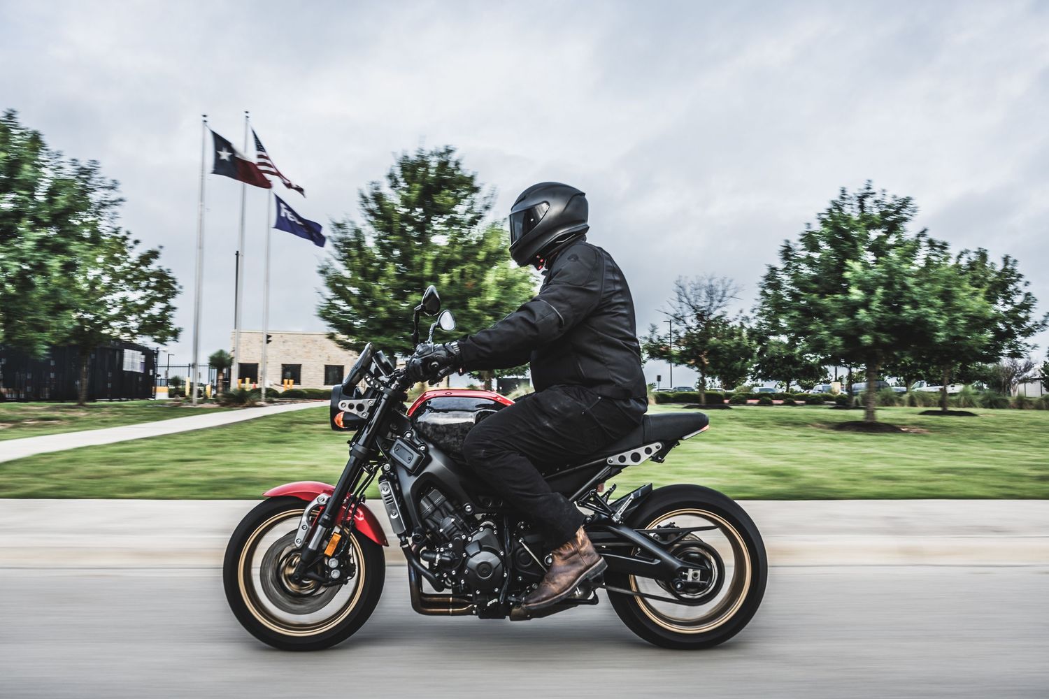 Motorcyclist riding black street bike past commercial buildings and American flag.