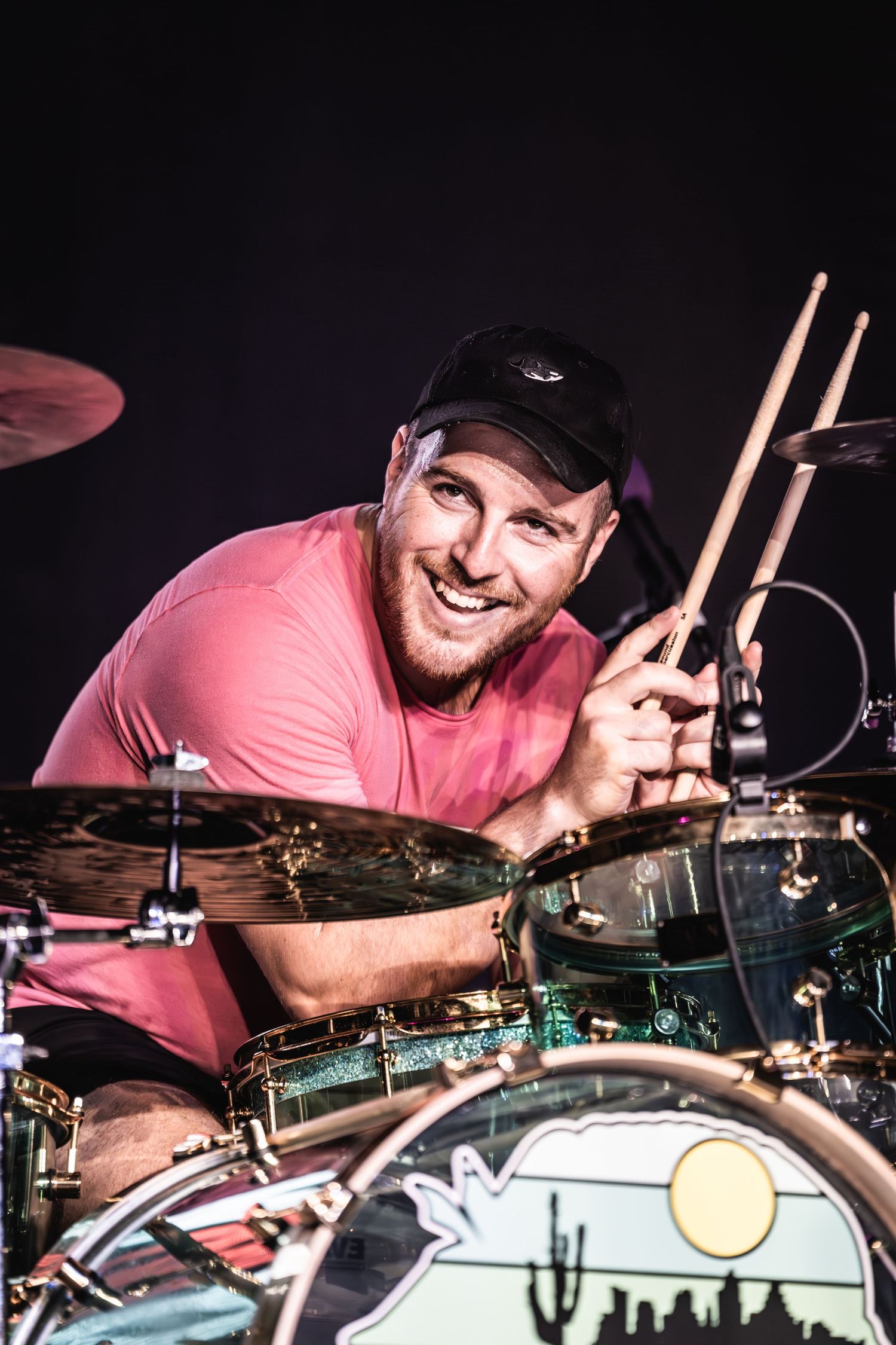 A drummer in a pink shirt smiles while holding drumsticks behind a drum kit with a desert-themed bass drum head.