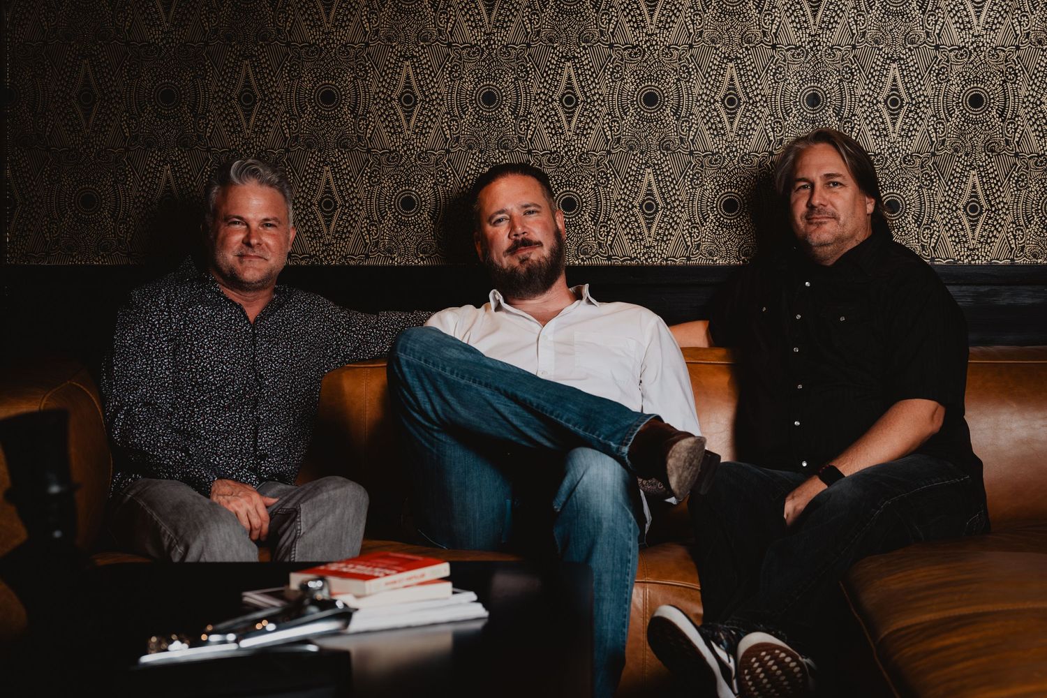 Three men seated on brown leather couch during professional photoshoot.