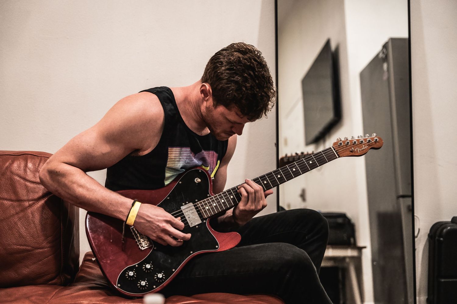 Musician playing electric guitar while sitting on brown leather couch in practice space.