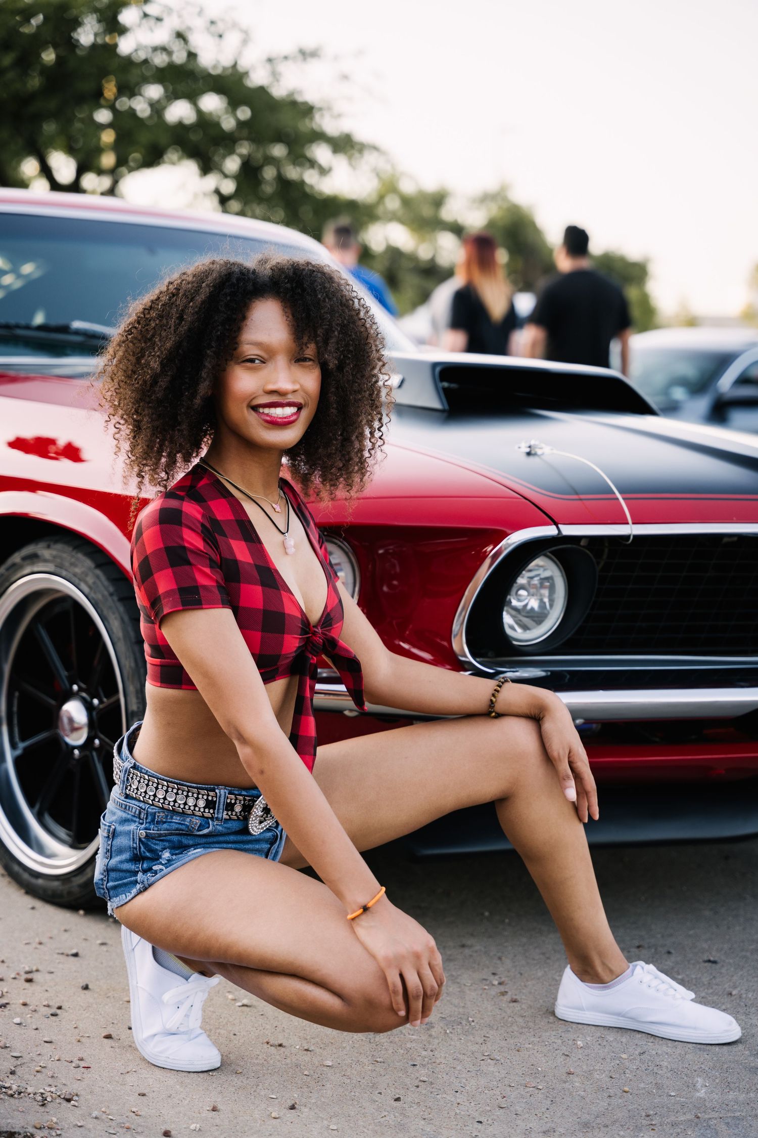Model in red plaid top and denim shorts poses beside vintage red muscle car.