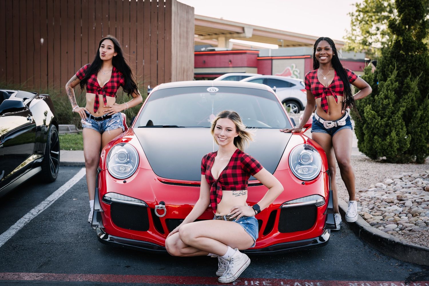 Three models in matching plaid tops pose with a red Porsche sports car.