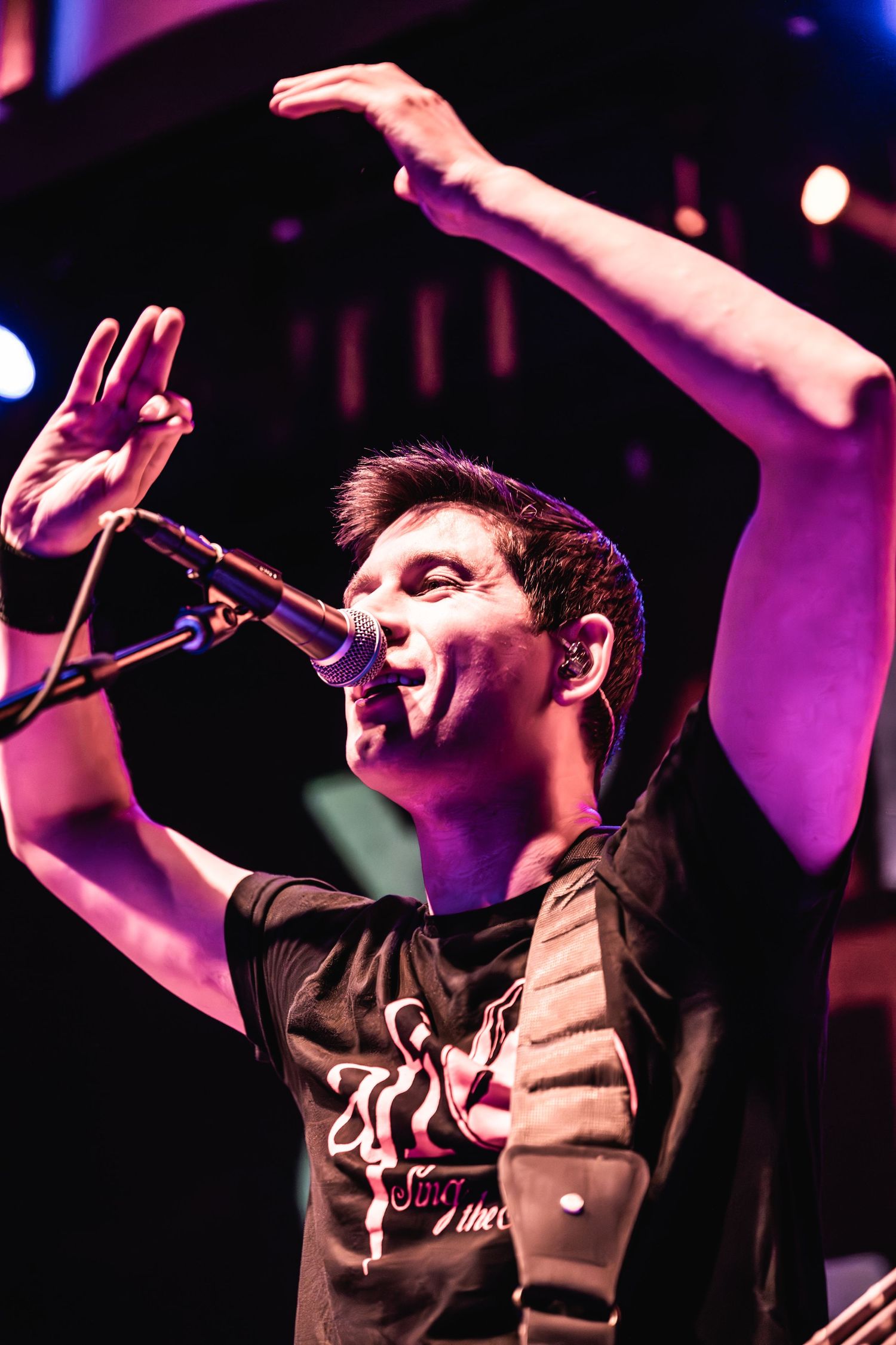 Performer raises arms while singing into microphone on darkened stage with dramatic lighting.