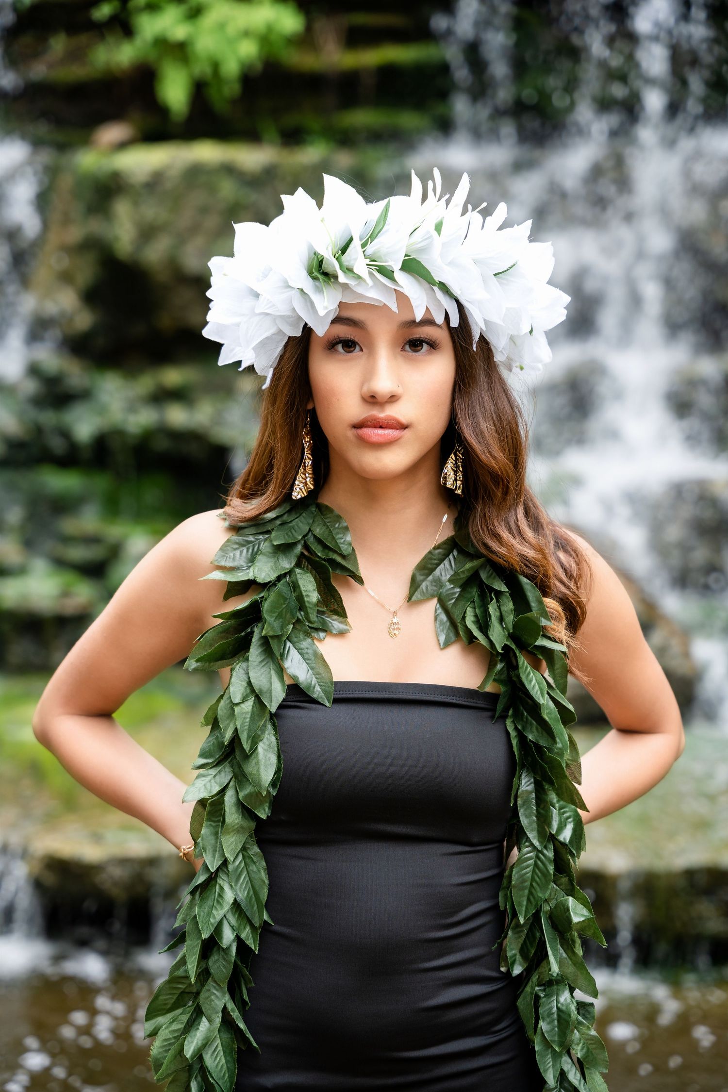 Graduate in black dress wearing white flower crown and green leaf lei poses in front of waterfall.