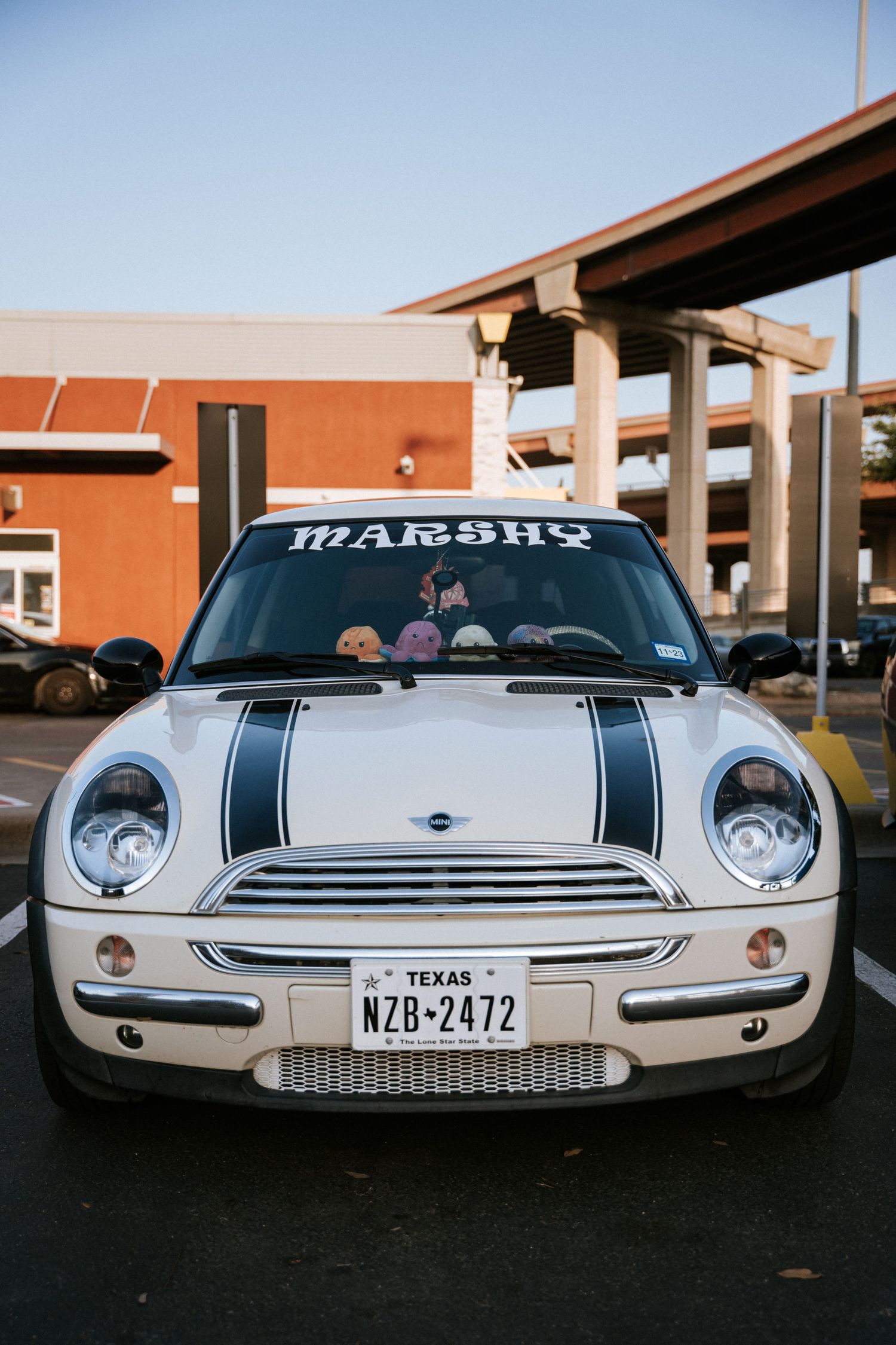 A white Mini Cooper with racing stripes and Texas plates parked in front of an orange building.