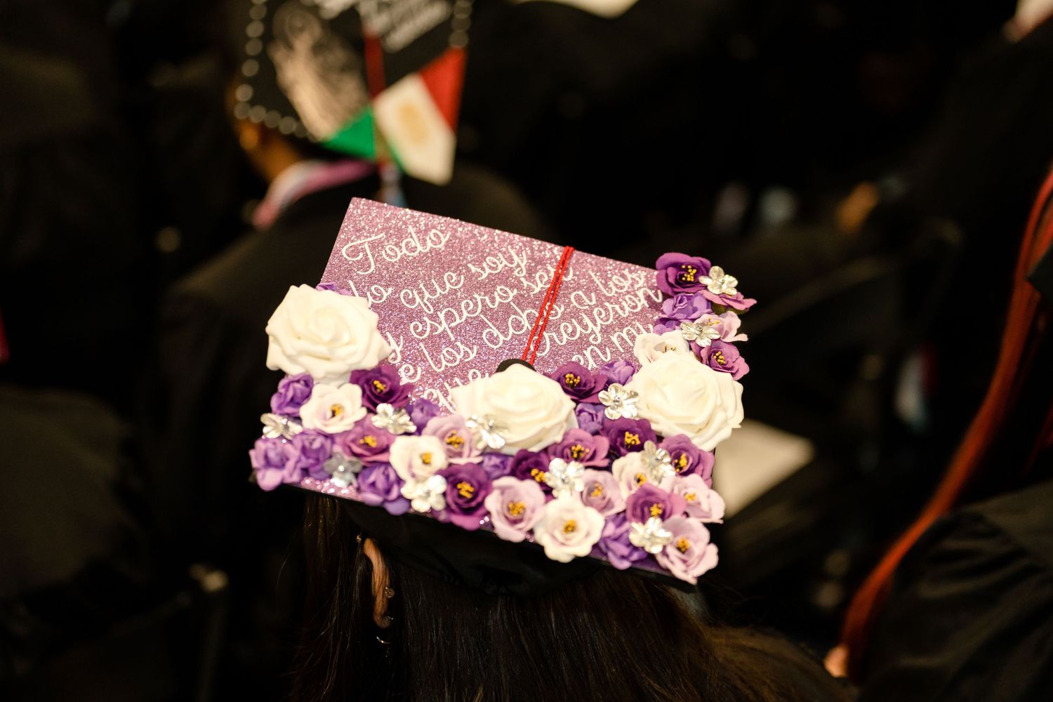 A graduation cap decorated with purple flowers and white roses on a dark background.