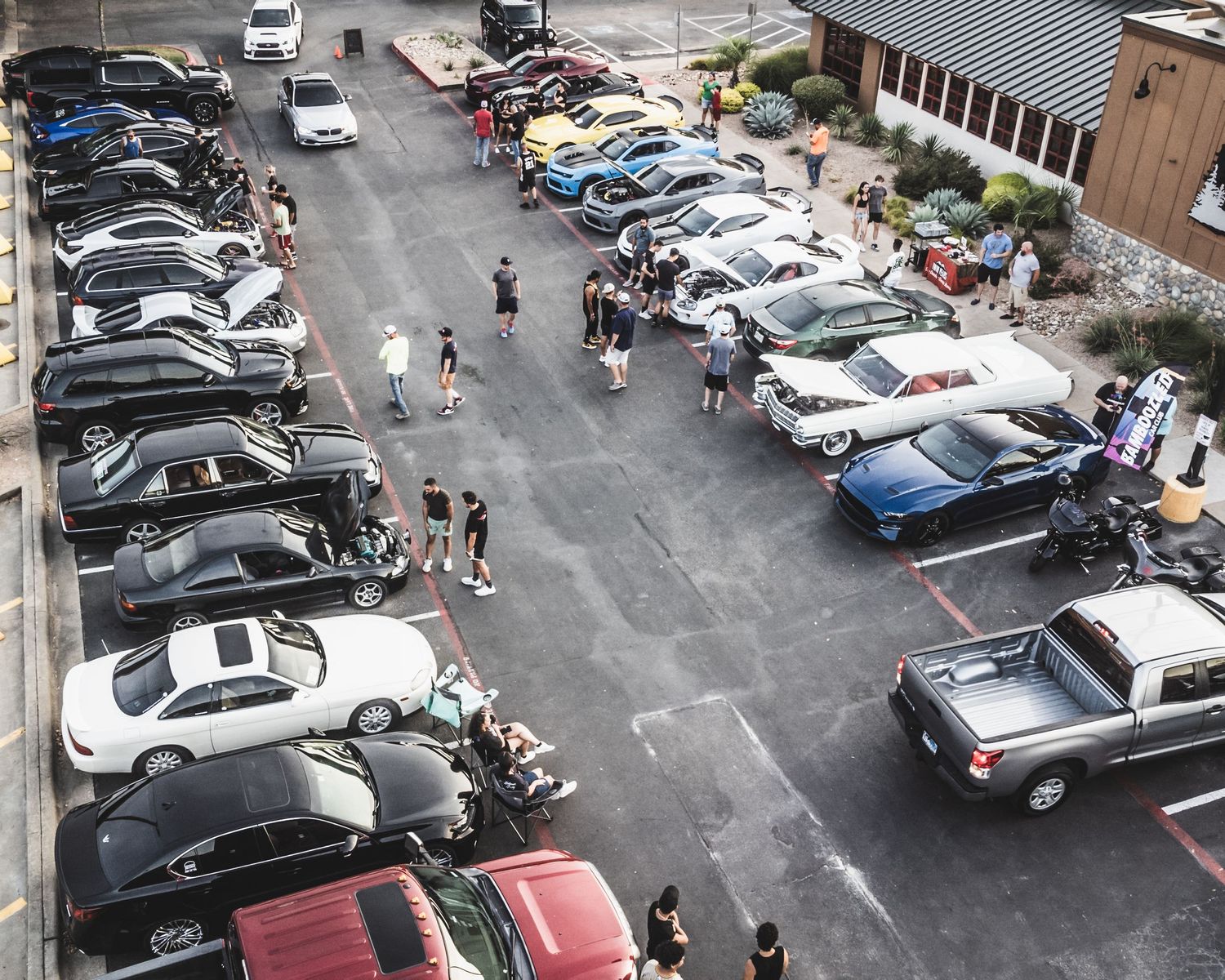 Aerial view of a car meet in a parking lot showing rows of parked vehicles and attendees gathering between them.