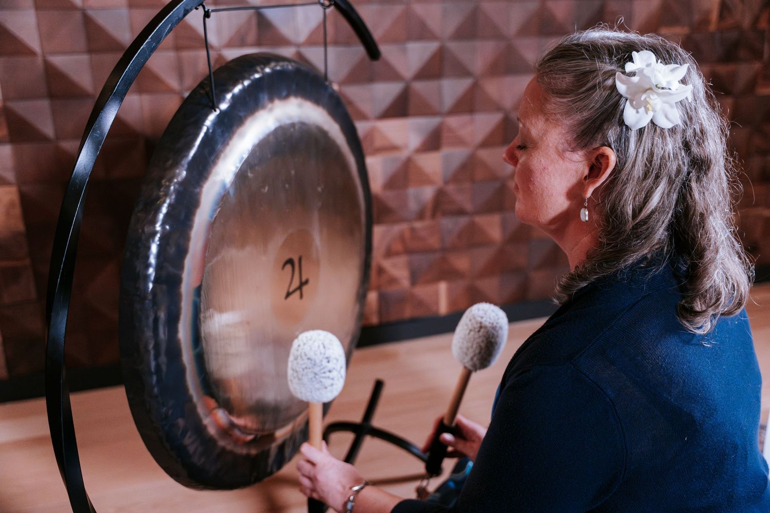 A performer plays a large gong instrument in a room with textured wall panels.
