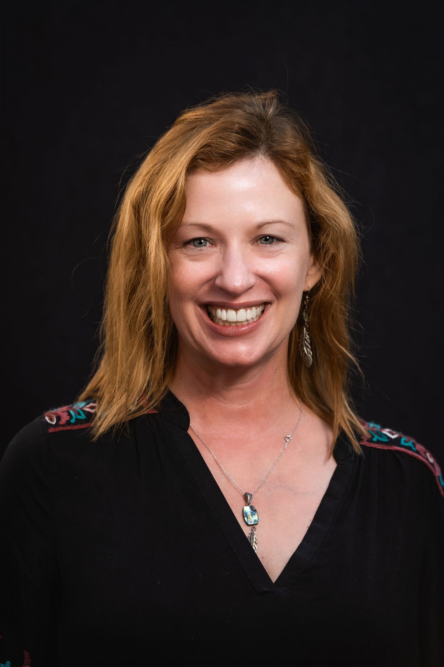Cheerful studio portrait with person wearing black top and pendant necklace against dark background.