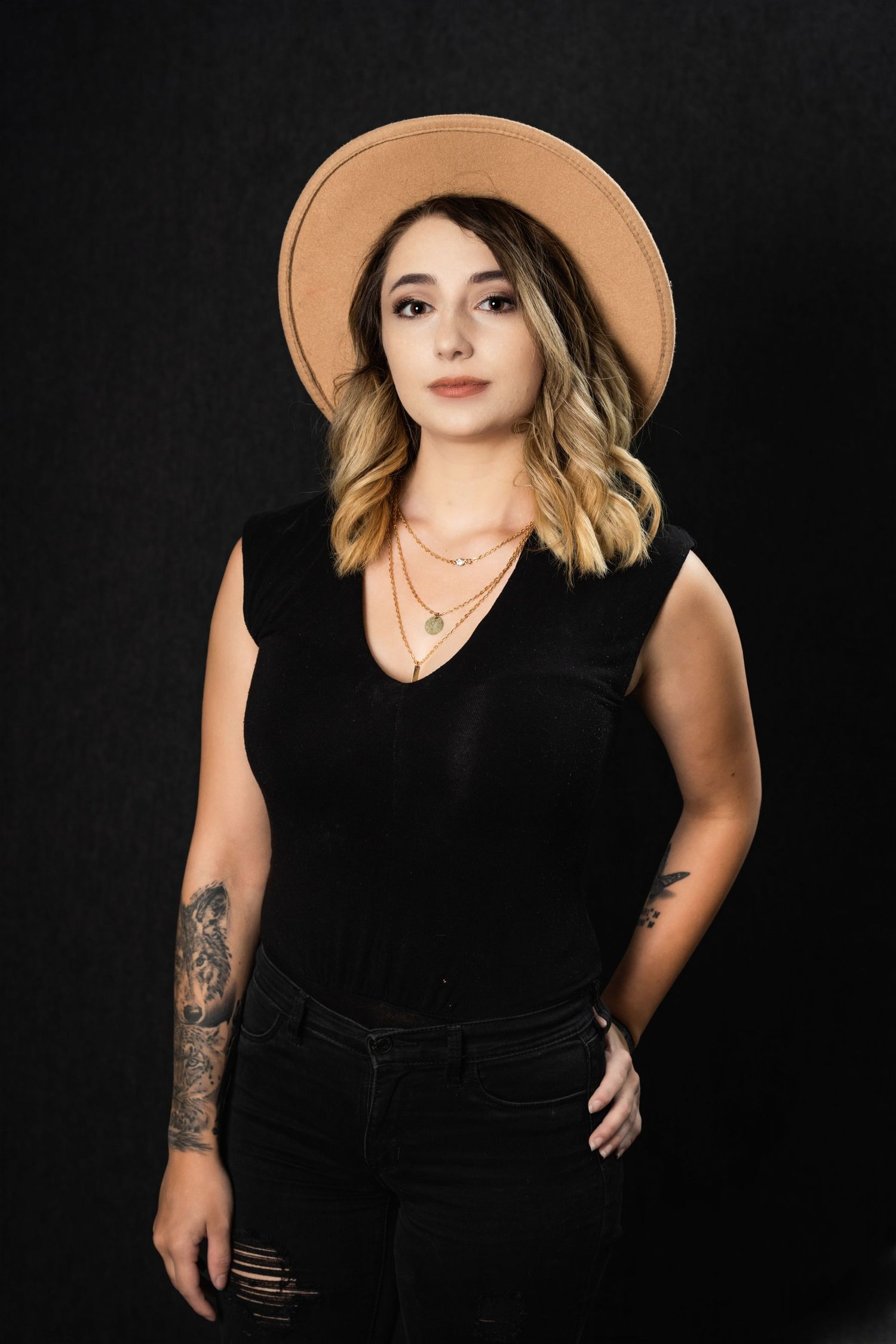 Modern portrait in black outfit with tan hat and visible arm tattoo against dark background.